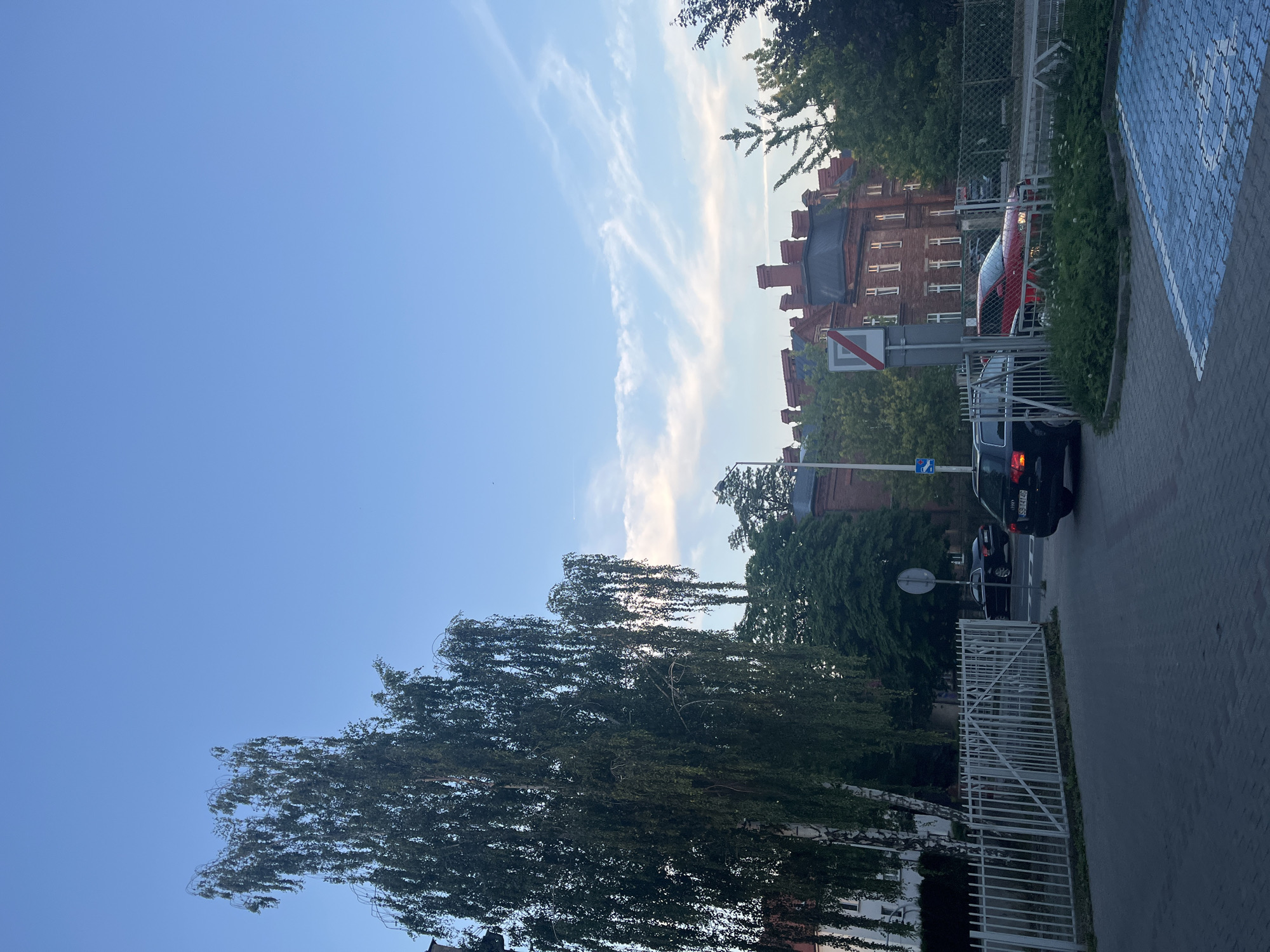 The image depicts a street scene in an urban environment under a bright blue sky with scattered wispy clouds. In the foreground, a paved area with blue-marked parking spots is visible. A dark-colored sedan is parked on the street, and a red sedan is partially visible behind a fence.
To the left, a tall birch tree with drooping branches stands prominently. Behind the tree, a white metal fence encloses an area with more greenery. Further in the background, a large, old brick building with multiple windows and a dark roof is situated. The building has several chimneys.
The street appears to be an alley or a parking lot entrance, with a road leading away from the viewer. Trees and foliage are interspersed throughout the scene, suggesting a moderately green urban setting. There are no people clearly visible in the image. The lighting suggests it is either early morning or late afternoon. The license plate of the dark sedan is partially visible and reads "GS 14146".