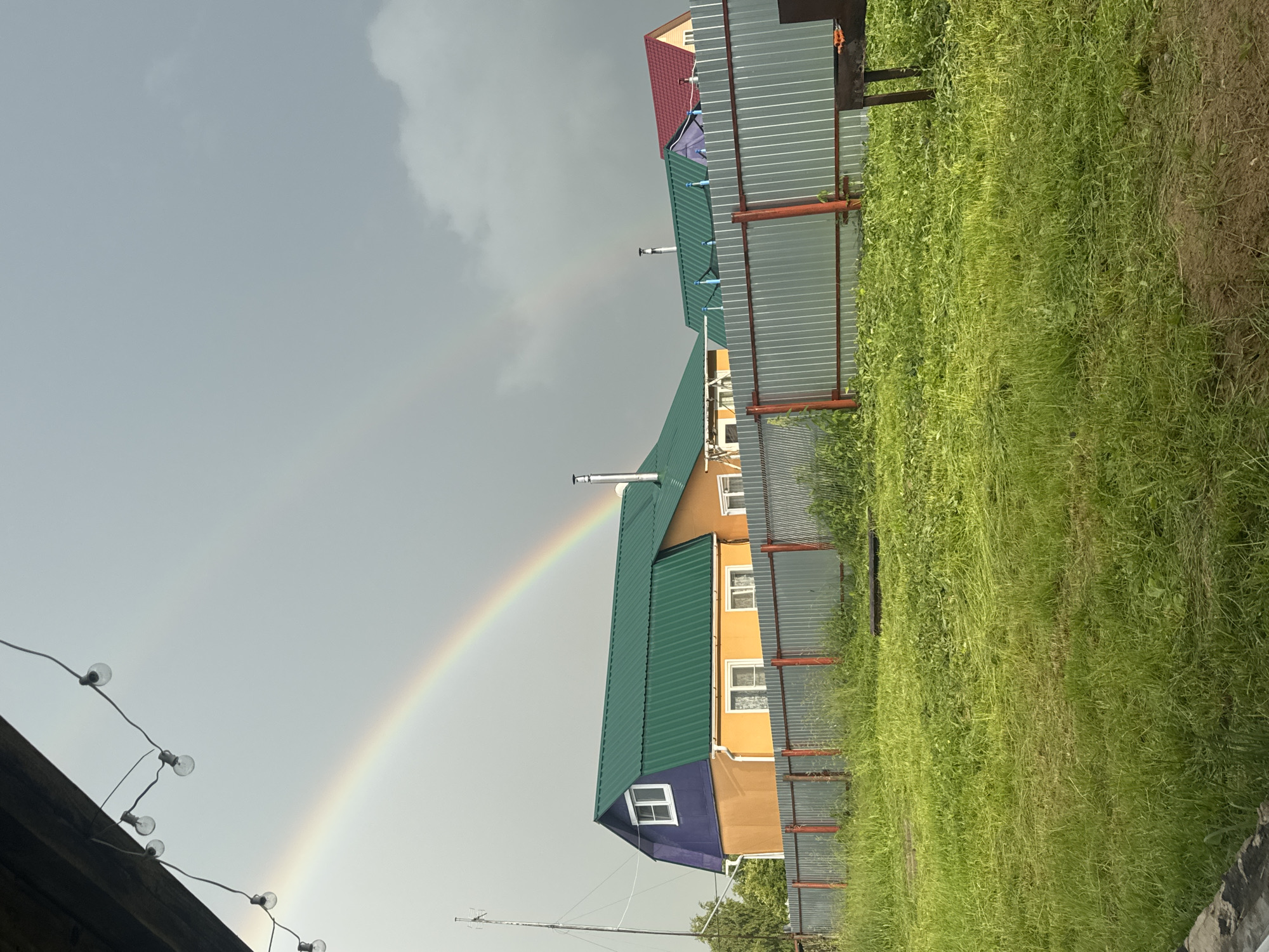 This is a vertical, eye-level, outdoor shot of a rural scene in Южное Снт, Russia. The weather appears to be overcast with a faint rainbow arching across the sky from left to right. The foreground is dominated by a lush, green grassy area, partially mown, with some patches of bare earth. A corrugated metal fence with red posts runs along the right side of the frame, separating the grassy area from the buildings.
Several houses with colorful, corrugated metal roofs are visible. The largest house in the center has a bright orange lower section and a bright green roof, with visible chimneys. A smaller section of this house is painted purple with a single white-framed window. Further back, another house features a red roof. The scene suggests a residential area in a dacha (summer house) community.
In the upper left corner, a string of incandescent light bulbs hangs from what appears to be a wooden structure, adding a decorative touch to the scene. There is no visible text in the image. The overall impression is one of a post-rain, slightly gloomy but picturesque day in the Russian countryside.