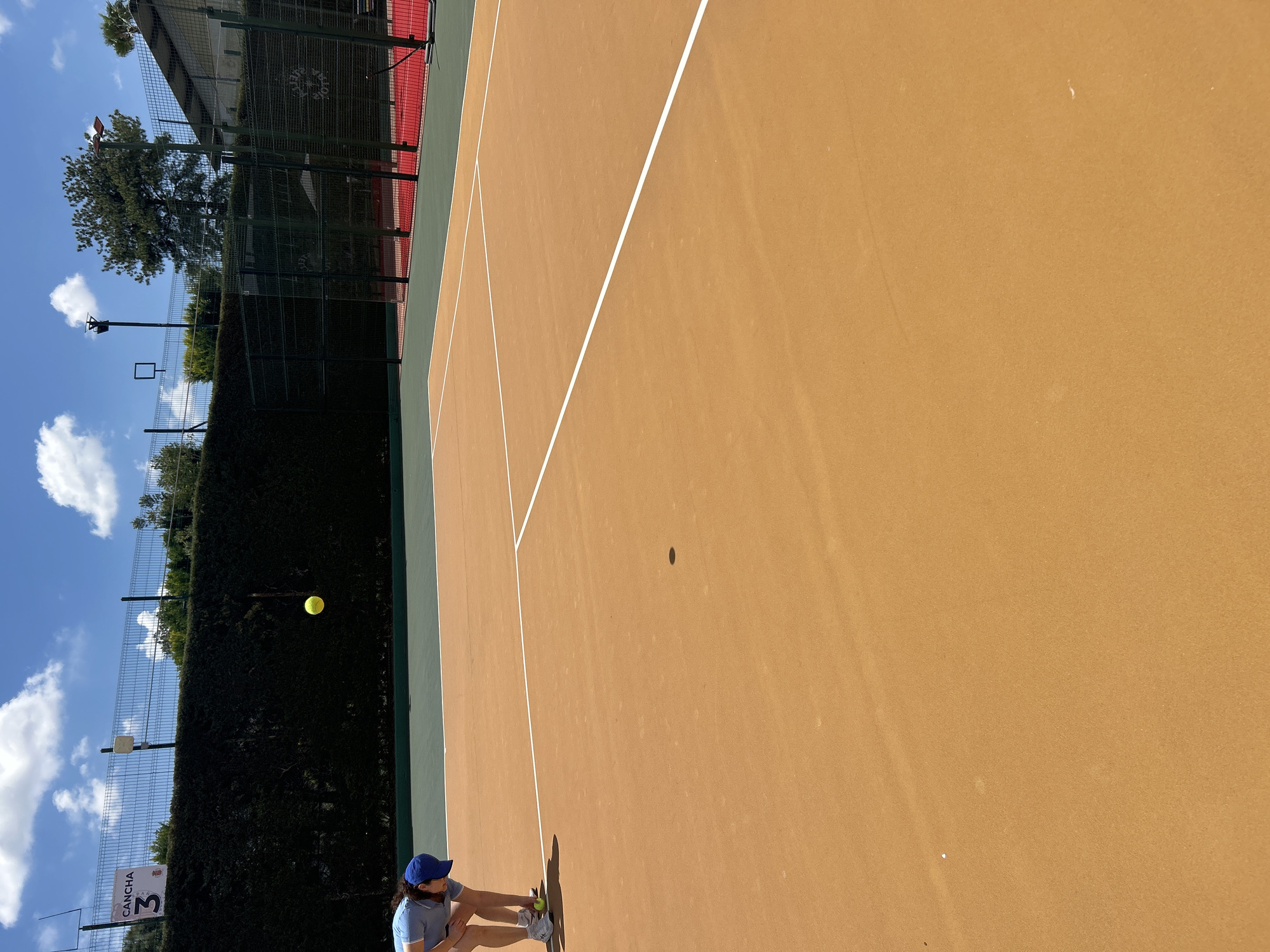 A ground-level, outdoor shot, rotated counterclockwise, depicts a tennis court in Puebla, Mexico. The court is tan-colored with white lines, partially visible. A woman in a blue hat and light blue shirt is seen in the lower left corner, seated on the ground with a tennis ball. A yellow tennis ball is suspended in the air near the court's boundary. A dark green hedge runs along the back of the court, with a chain-link fence and trees visible beyond. A sign reading "CANCHA 3" is affixed to the fence. The sky, a mix of blue and white clouds, is visible above the hedge. Buildings and additional trees are in the background.