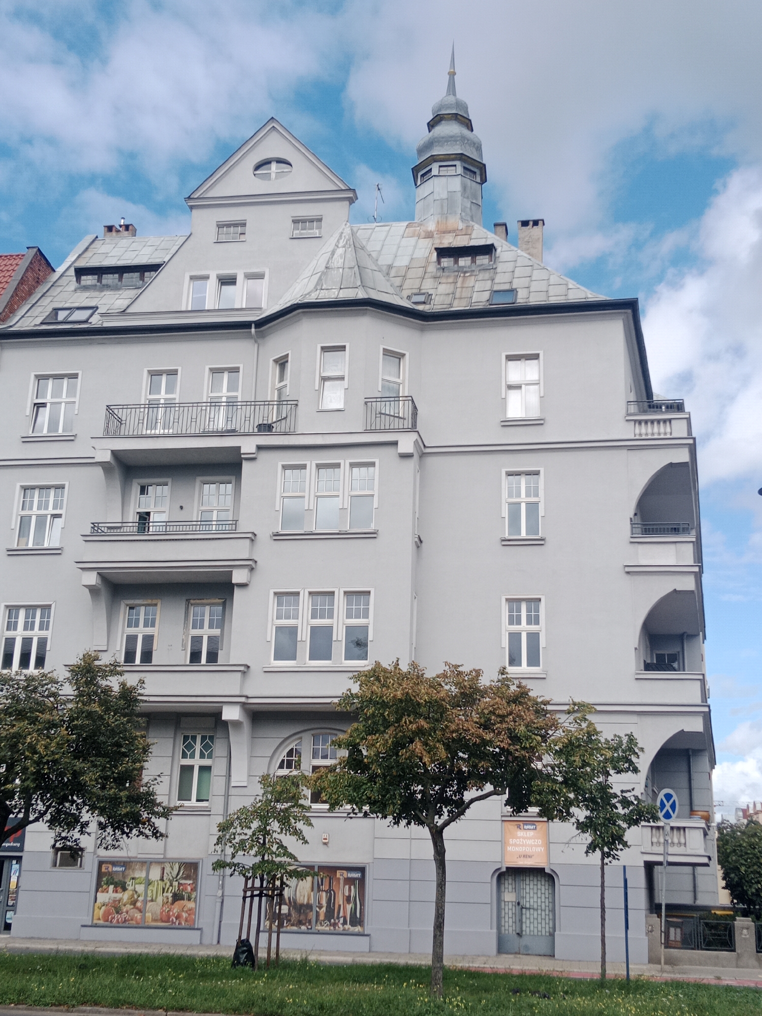 This is a photograph of a multi-story building with a gray facade, located at Plac Wolności 1 in Bydgoszcz, Poland. The building has a distinctive architectural style with a tower topped by a spire, a pitched roof with dormer windows, and several balconies. The facade is punctuated by numerous white-framed windows of various sizes. The ground floor appears to be commercial, with large display windows featuring images of produce and wine. Trees with green and reddish-brown foliage are planted in front of the building, along with a patch of green grass and some yellow wildflowers. The sky above is blue with scattered white clouds. A blue sign with a white cross symbol is visible to the right of the building.