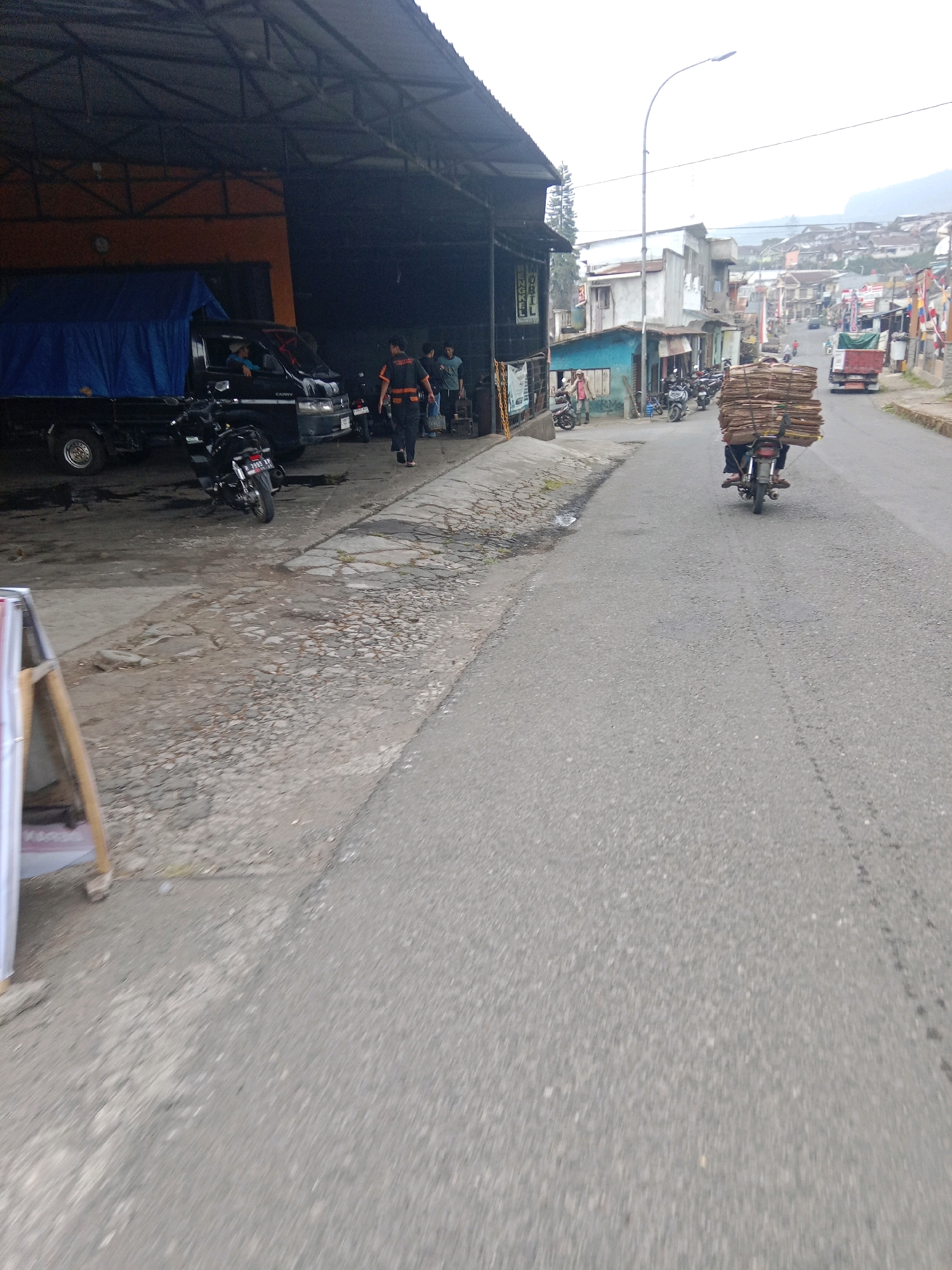 A street scene in Bujungsari, Indonesia shows a paved road with a slight incline. On the left, a dark-colored truck with a blue tarp is parked under a large open-air shelter. Several motorcycles are parked in front of and beside the truck. Three men are walking away from the shelter towards the road. The man in the foreground is wearing a dark shirt with orange accents and dark pants.
On the right side of the road, a person is riding a motorcycle loaded with a large stack of cardboard. The motorcycle is facing away from the camera, heading up the incline. Buildings of varying heights and styles line the right side of the road, some appearing to be residential homes and others possibly small businesses. The sky is overcast, contributing to the muted lighting of the scene. Further up the road, more buildings are visible on a hillside, with a truck also seen further in the distance. A street lamp is positioned on the side of the road.