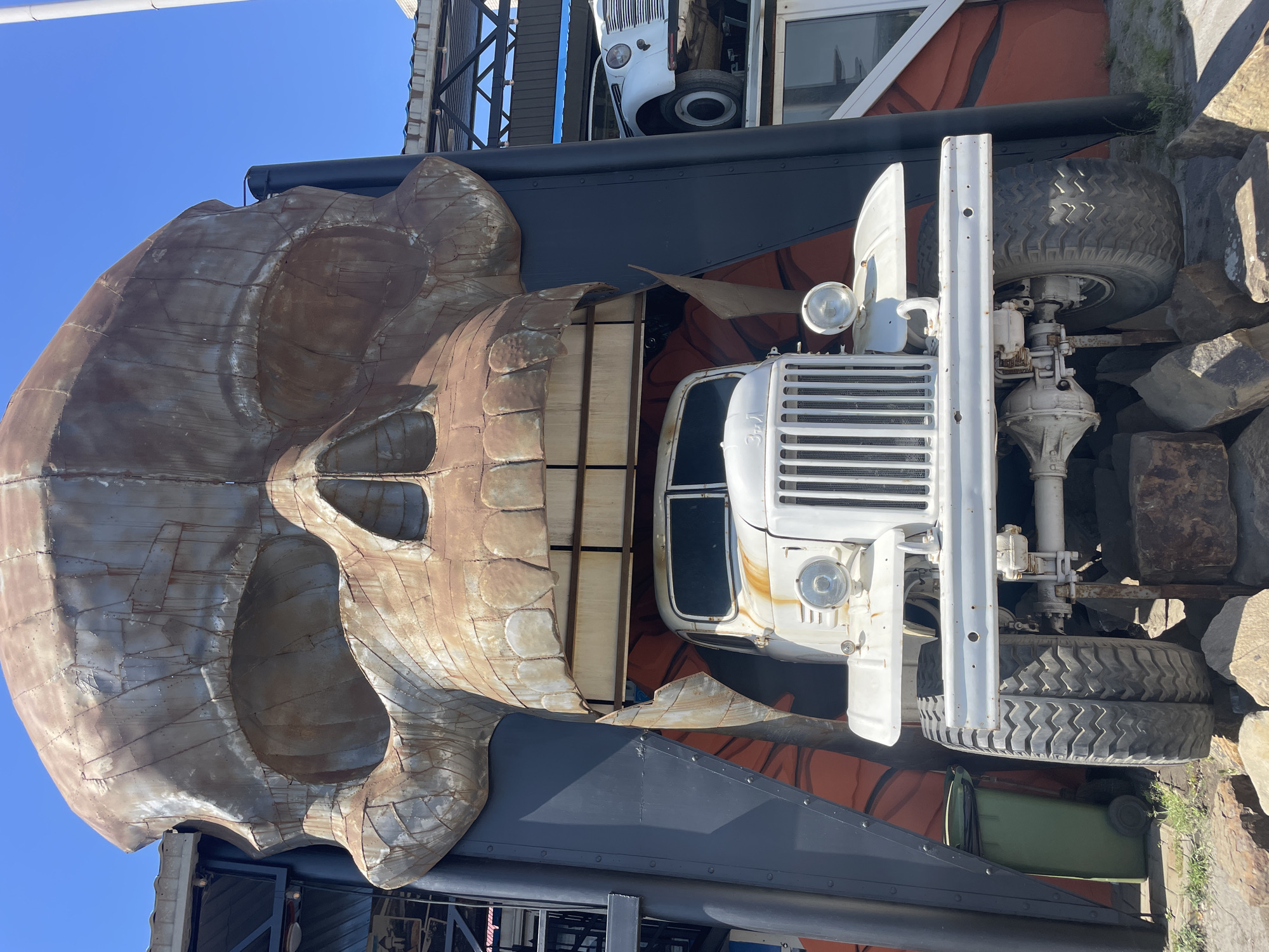 The image depicts a large, weathered, metal sculpture of a skull with its mouth open, framing a white truck. The skull is constructed from various pieces of metal, showing signs of rust and wear. The truck appears to be an older model, possibly a Soviet-era vehicle, with a prominent grille and large tires. It is positioned within the skull's maw, supported by metal beams and rocks. In the background, parts of a building are visible, including a corrugated roof structure and another white vehicle parked on a higher level. A green trash bin is situated to the left of the skull. The scene is set outdoors under a clear blue sky. This sculpture is part of the Alexander Burenkov's Museum of the Art of Mechanical Sculptures located in Kamensk-Shakhtinsky, Russia. There are no people visible in the image.