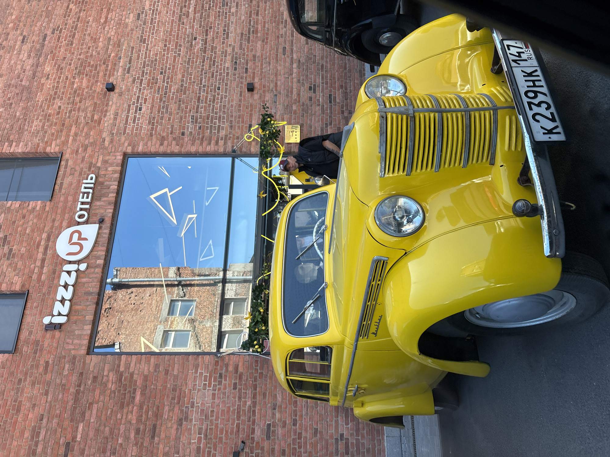 The image shows a vintage yellow car parked in front of a brick building with a large window. The building has a sign that reads "izzzi UP ОТЕЛ". The car is a GAZ-21 Volga. There is a man standing next to the car, wearing a black vest and a red baseball cap. Another vintage car, a black GAZ-21 Volga, is partially visible to the right of the yellow car. The scene appears to be in Saint Petersburg, Russia, as indicated by the car's license plate which reads "K239HK 147 RUS". The building's architecture and the car models suggest a historical setting. The yellow car is the primary subject of the image, with its gleaming paint and chrome details prominently displayed. The man standing by the car seems to be attending to it or posing for a photo. The reflection in the window shows the building opposite and the sky.
