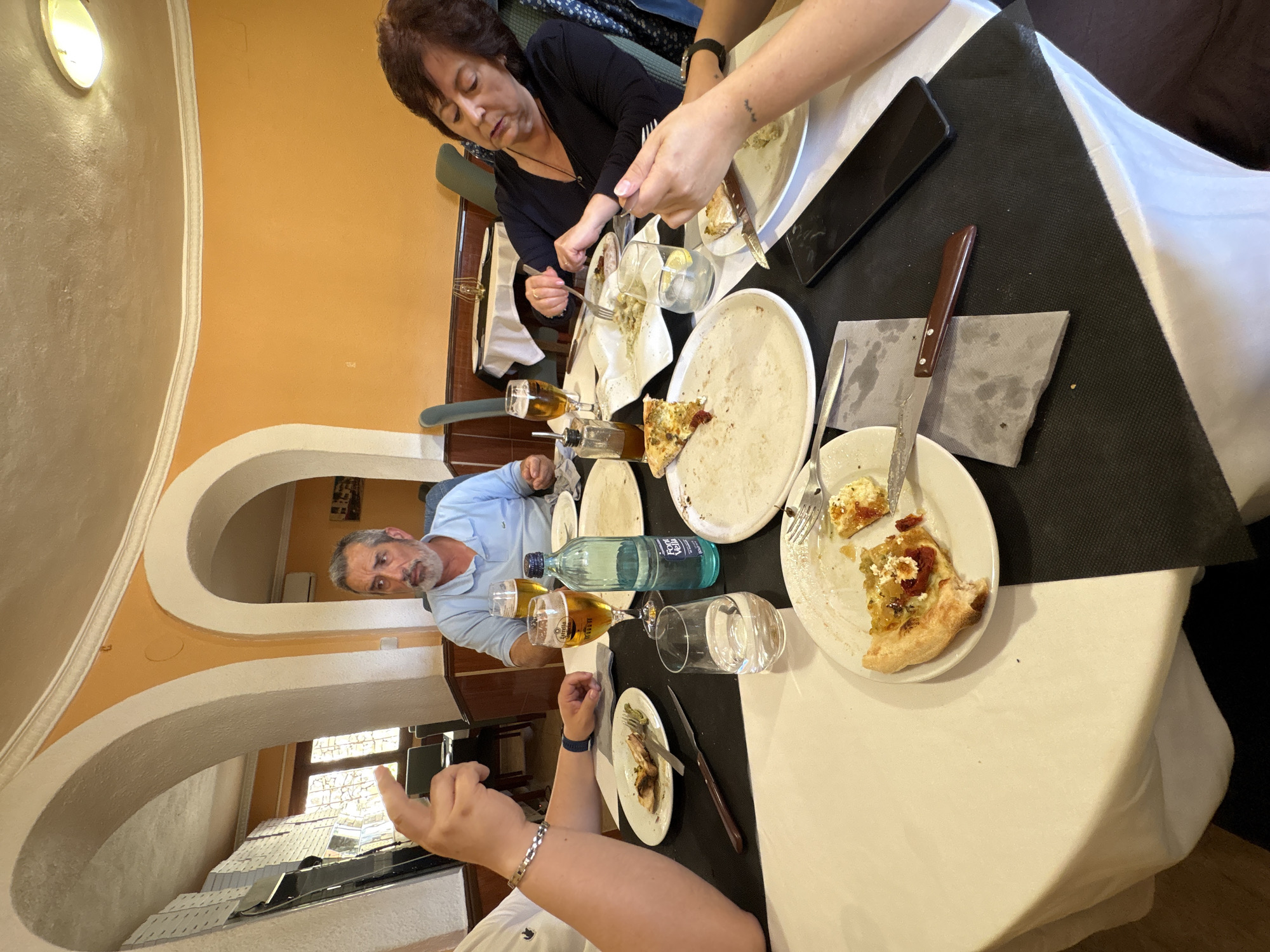 The image shows a group of people dining at a table. The table is covered with a black and white tablecloth and has plates with leftover food, silverware, glasses, and a water bottle on it. There are at least three adults present, with one middle aged woman seated. Another man is seen in the bottom left, and an individual on the right has a small tattoo. The setting appears to be inside a restaurant with arched walls and a view of the city visible through a window. The location could be Teulada, Spain.