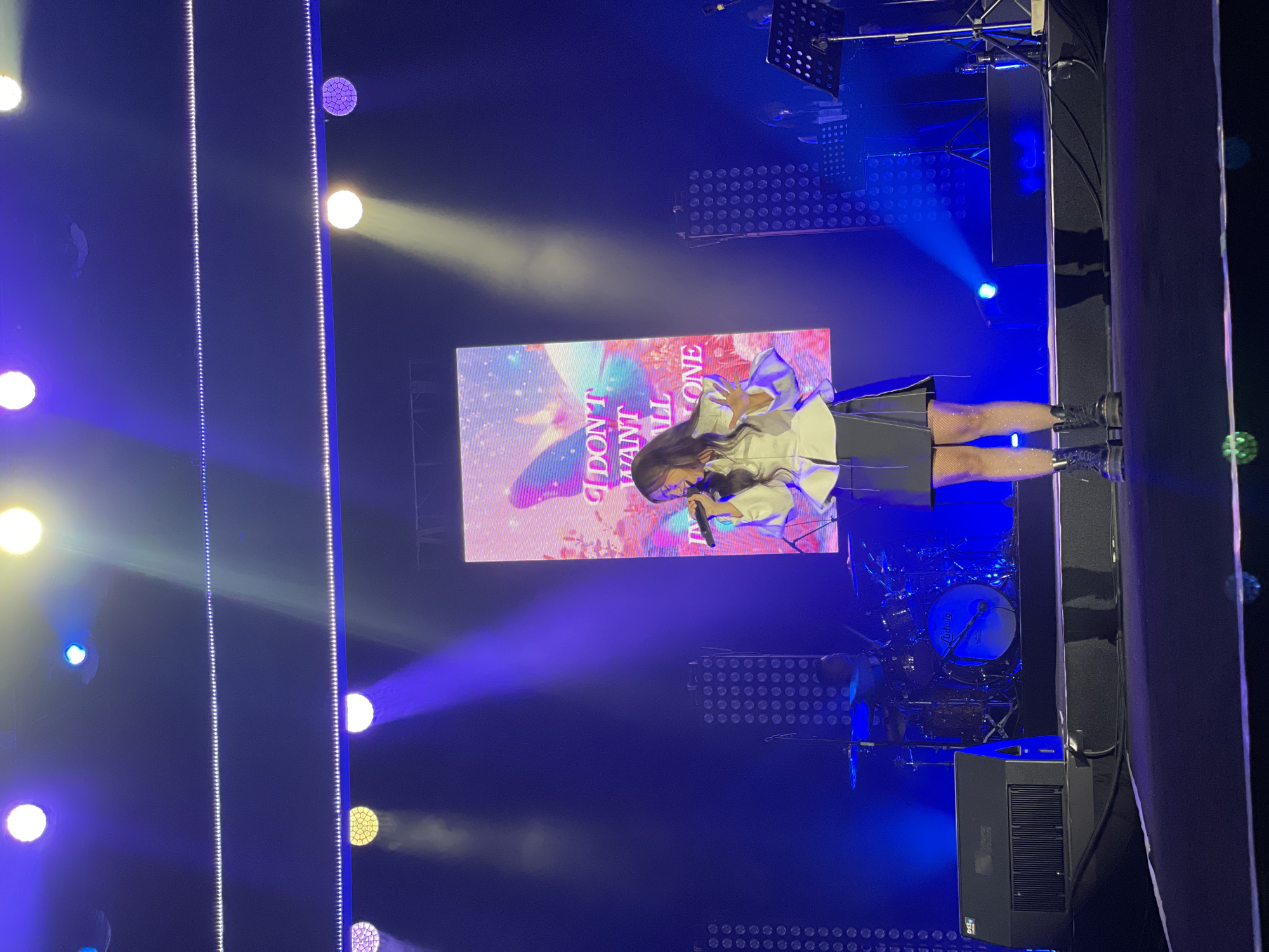 A woman is singing on a stage in Salembaran, Indonesia, illuminated by stage lighting. A large screen behind her displays a graphic with the text "I DON'T WANT ALL ONE." Musical equipment, including drums and speakers, is visible on the stage. The stage is lined with lighting fixtures.