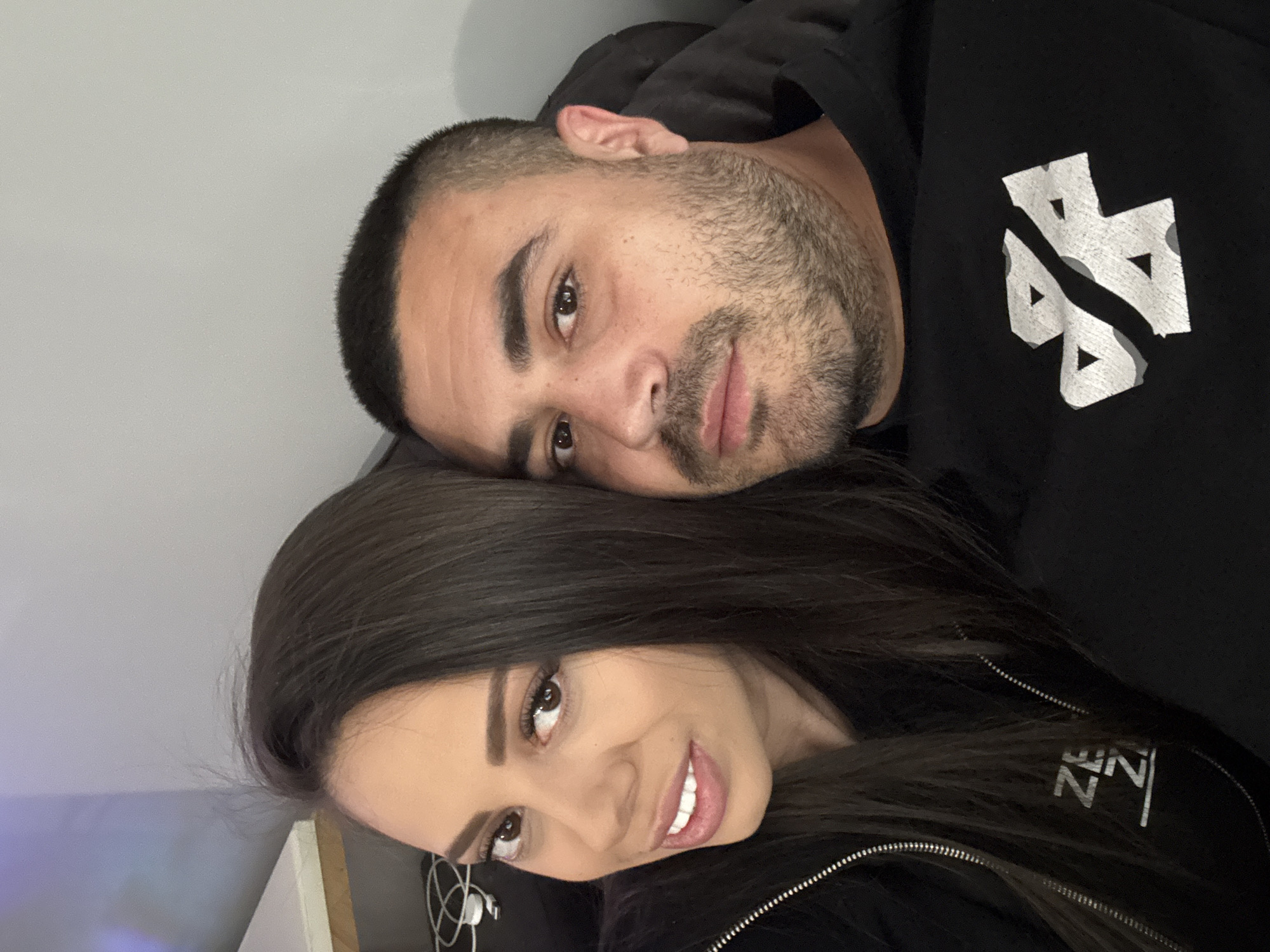 A close-up, informal portrait features a man and a woman, seemingly a couple, positioned intimately. The image is oriented sideways, with the subjects lying down or leaning.
The woman is in the foreground, facing the camera with a warm, genuine smile. She has long, dark brown hair, which frames her face and flows over her shoulders. Her eyes are dark brown, and she has well-defined eyebrows. She is wearing a black zippered jacket or hoodie, and part of a white logo or text is visible on it, partially obscured by her hair, appearing to spell "ZEN" with a stylized 'Z'. White earbud wires or cables are faintly visible near her left ear.
Behind and slightly above her, the man rests his head on her, looking towards the camera. He has short, dark hair, closely cropped on the sides, and a dark beard covering his jawline and chin. His expression is soft and content, with a slight smile. He is wearing a black hooded sweatshirt with a prominent white, abstract logo on the chest. The logo consists of two interconnected, stylized shapes, possibly resembling a split '44' or a distorted percentage sign, with small dotted textures within its forms.
The background is a simple, light grey wall, appearing out of focus, suggesting an indoor setting like a home. The overall mood conveyed is one of affection, happiness, and relaxation. The image captures a personal moment between the two individuals. While the context specifies Varna, Bulgaria, there are no visual cues within the image itself to confirm this location.