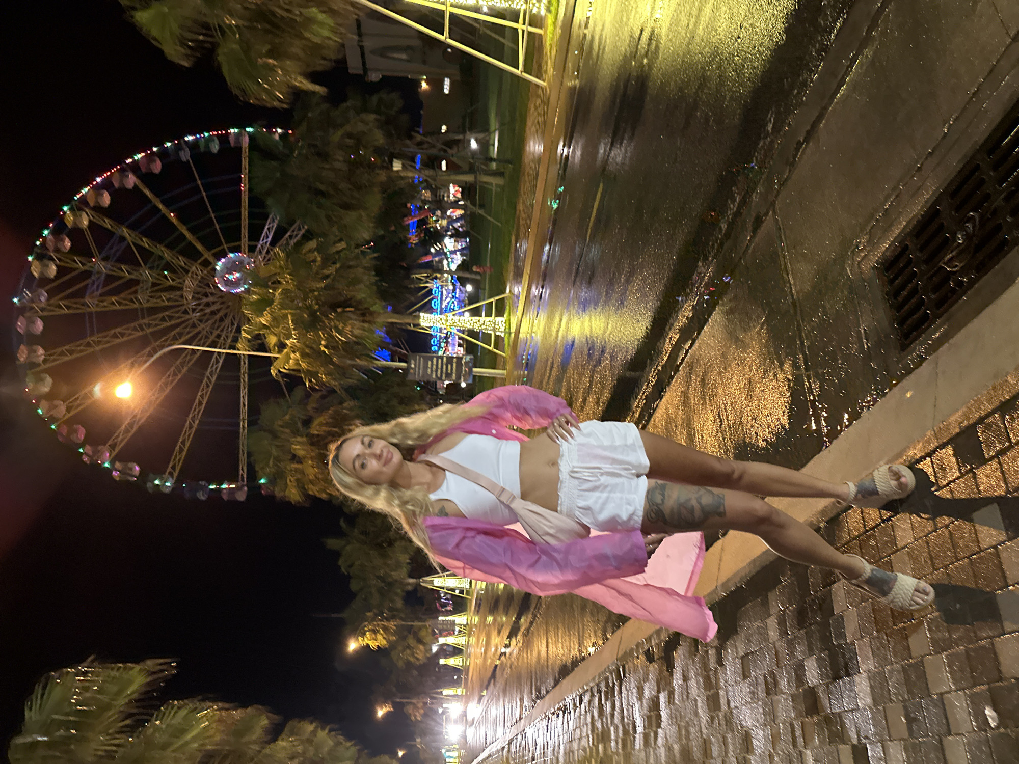 The image displays a vibrant nighttime scene in an outdoor entertainment area, likely a promenade or boardwalk, in Tien Thanh Ward, Vietnam. The ground is visibly wet and reflective, suggesting recent rain or irrigation.
In the center of the frame, a young woman with long blonde hair stands, posing for the camera. She is dressed in a white crop top, white shorts, and a sheer, flowing pink long-sleeved jacket. She wears light-colored sandals and has tattoos visible on her legs. A light-colored cross-body bag is slung over her shoulder. Her expression is neutral as she looks directly at the viewer.
To the left, a large Ferris wheel dominates the background, brightly lit with colorful lights along its rim, including red, green, and blue, and a prominent, luminous center. Several palm trees are scattered throughout the scene, some partially obscuring the Ferris wheel and others lining what appears to be a street or pathway. Further in the background, other bright artificial lights are visible, contributing to the illuminated atmosphere.
The wet, paved surface in the foreground and to the right reflects these lights, creating streaks of color and glow. A dark metal drainage grate is embedded in the wet pavement on the right side of the image. The overall impression is one of a lively evening at a tropical or sub-tropical resort or amusement area. No discernible text is visible in the image.