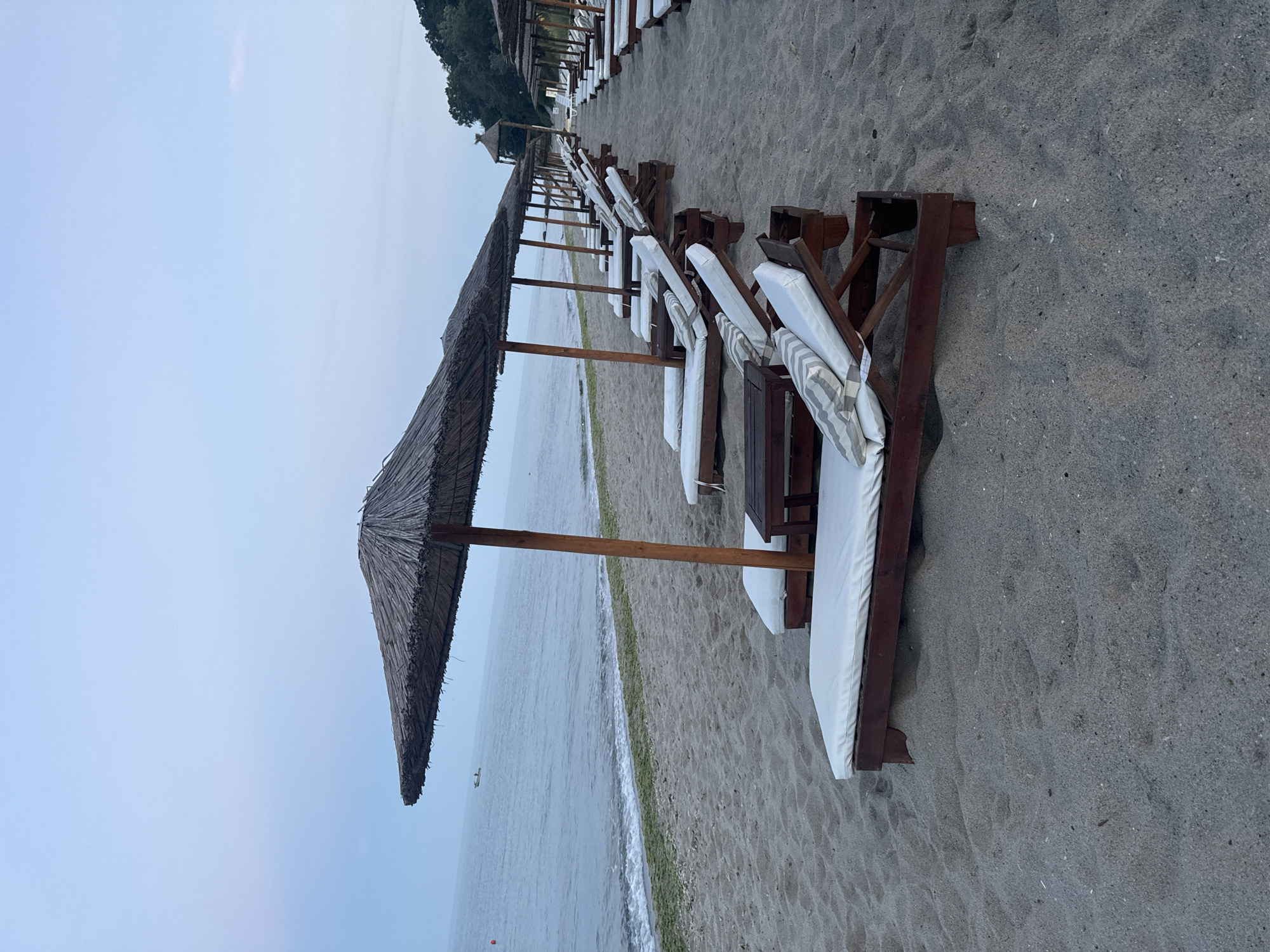 This image depicts a row of empty sun loungers with straw umbrellas on a sandy beach. The sun loungers are made of dark wood with white cushions and some have striped pillows. The beach is situated along the Black Sea coast, with calm, gray water visible in the background. A small boat is present in the distance on the water. The sky is overcast with a pale blue hue. The overall scene suggests an early morning or late evening atmosphere on a quiet beach, likely a resort area in Varna, Bulgaria. No people are present in the image. The environment is a typical beach setup with loungers arranged for relaxation.