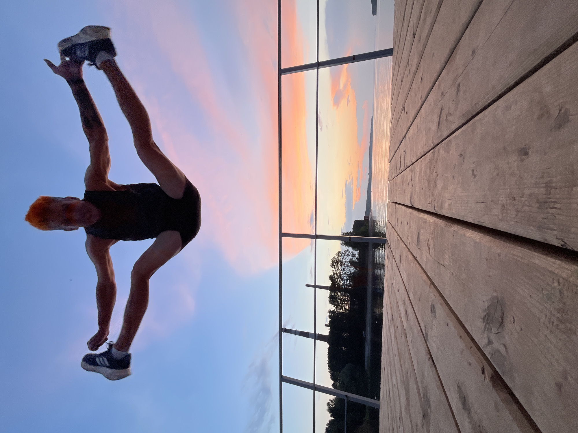 In the city of Chaikovsky, Russia, at sunset, a man with vibrant orange hair is captured mid-air performing a split jump. He is wearing a dark tank top and black shorts. His arms are outstretched to his sides, and his legs are extended horizontally, forming a perfect split. The man is positioned above a wooden pier, which occupies the foreground of the image. Beyond the pier, a body of water is visible, reflecting the soft hues of the sunset. In the background, across the water, there is a tree line, and two industrial chimneys stand tall. The sky is a canvas of pastel colors, with shades of blue, pink, and orange blending together. A metal railing separates the pier from the water and the distant landscape.