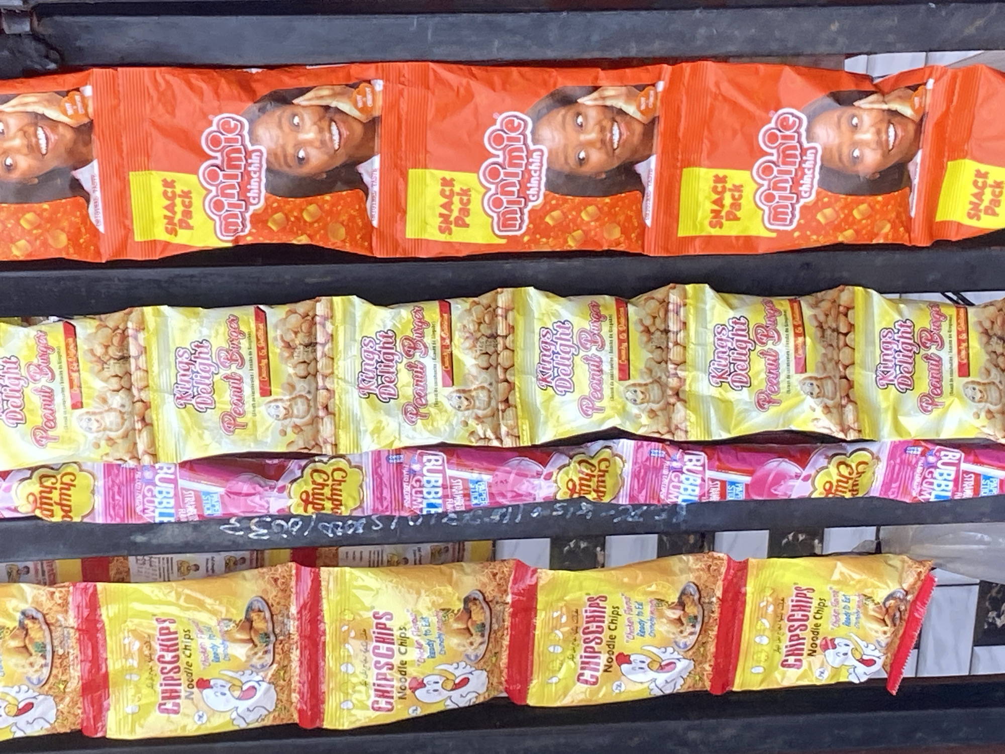 The image presents a close-up view of multiple rows of snack packages displayed on a retail shelf or stall, likely in Sasaro, Nigeria.
The top row features several orange "minimie chinchin SNACK Pack" bags, each adorned with a smiling woman's face and showing pieces of chin chin.
Below this, the second row consists of yellow "Kings Delight Peanut Burger" packages, featuring a cartoon peanut character and marketing text like "Crunchy & Nutritious."
The third row displays pink and yellow "Chupa Chups BUBBLE GUM" lollipops, with "STRAWBERRY" flavor visible on some.
The bottom row showcases yellow "CHIPSCHIPS Noodle Chips" bags, which feature a cartoon chicken and text such as "Chicken Flavor" and "Ready to Eat Crunchy."
The bright, colorful packaging indicates items intended for sale. No specific activity is taking place other than the display of products. Some handwritten text, possibly numbers and letters like "15800010033" or pricing, is visible on a dark support beam between the rows, though it is partially obscured. The scene captures a typical assortment of snacks available in a local market or shop.