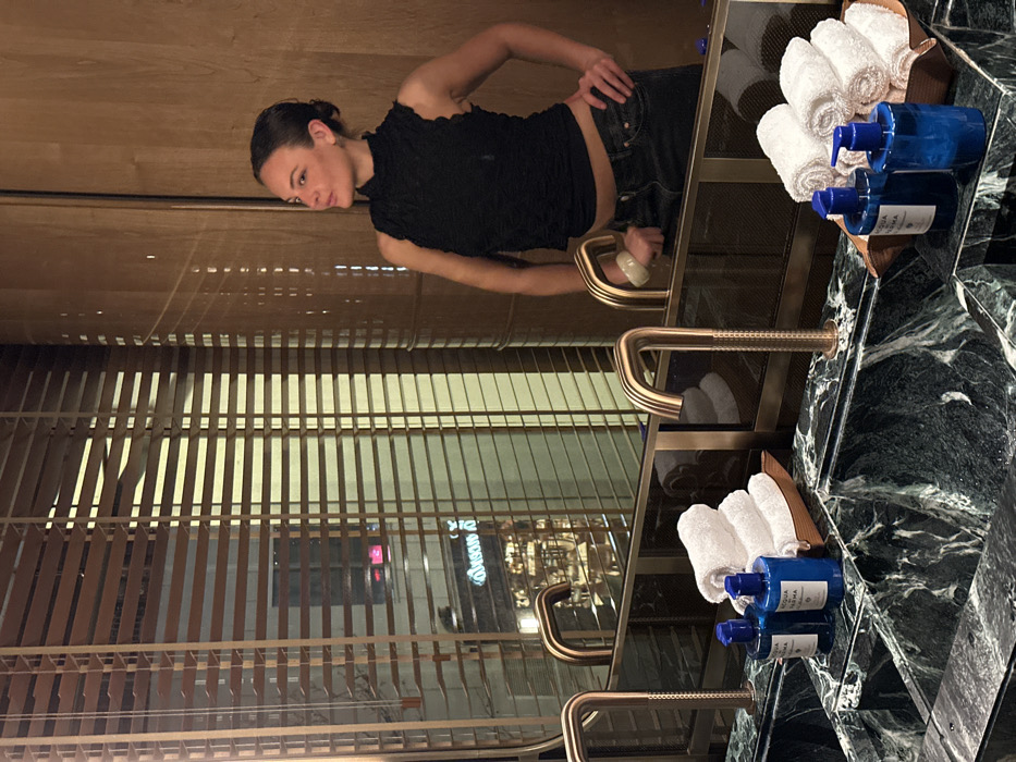 A person stands to the side of a bathroom vanity with marble counters. Adjacent to the counter is a mirror, which reflects a set of blinds, revealing a glimpse of the exterior. Soap and hand towels are neatly organized to the side of the vanity. The person's expression is thoughtful, with their hand resting on their hip.
