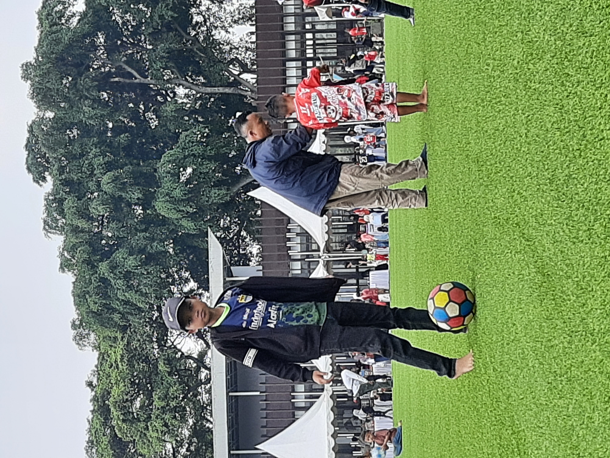 The image depicts a bright, overcast day on a vibrant green artificial turf field. In the foreground, a young boy wearing a dark hooded jacket over a blue and green jersey, dark jeans, and a baseball cap stands barefoot, looking towards the camera. A colorful soccer ball rests at his feet. To his right, a man in a blue jacket and beige pants is leaning down to interact with another young boy dressed in a red and white patterned outfit. In the background, several white tents are set up, with numerous people milling about. Behind the tents and people, a large building with a dark, patterned facade and several windows can be seen. Towering over the scene is a massive tree with dense green foliage. The setting appears to be an outdoor event, possibly a sports festival or community gathering, located in Bandung City, Indonesia.