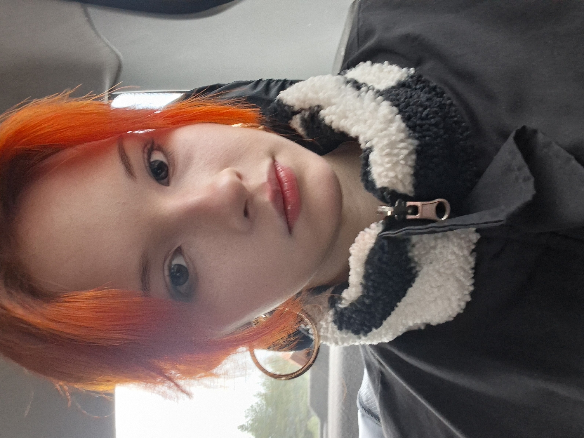 A selfie taken from a low angle shows a young woman with orange hair. She has light skin, light eyebrows, and dark eyes. Her lips are a reddish color. She wears a large gold hoop earring. The woman is wearing a dark gray jacket with a collar featuring a black and white striped pattern. A zipper is visible on the jacket. The background is blurry, with a glimpse of trees and daylight visible through a window. The location is possibly inside a vehicle, suggesting the city of Chaikovsky, Russia.