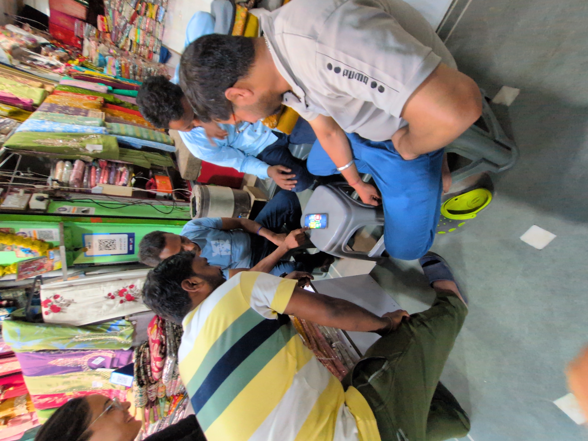 In a brightly lit store in Kullu, India, an array of colorful textiles hangs from racks and is stacked on shelves, creating a vibrant backdrop. Four individuals are gathered around a small grey stool. A man wearing a yellow, green, and white striped t-shirt and green pants is crouched down, looking at a smartphone on the stool. To his right, a man in a light blue shirt and dark pants sits on the floor, also focused on the phone. Another man, wearing a light blue long-sleeved shirt and dark pants, is seated on a stack of fabrics to the left, his gaze directed towards the phone. To the right, a man in a grey t-shirt with "Puma" branding on the sleeve, wearing blue pants, sits on the stool, looking down at the phone. A light blue slipper lies on the floor near him. The scene appears to be an informal gathering, possibly for shopping or social interaction, within a retail environment.