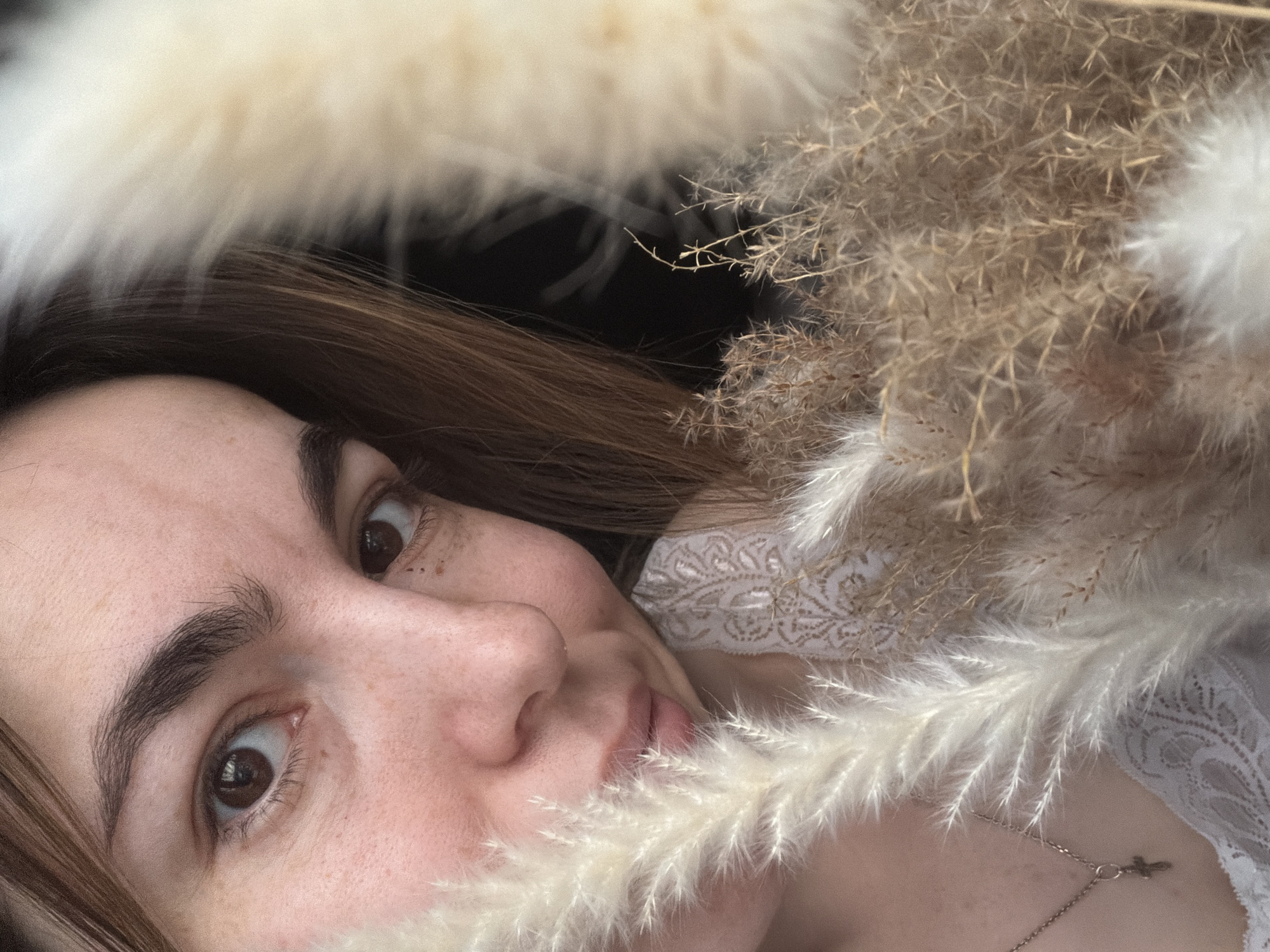 A close-up, angled shot shows a woman's face surrounded by dried floral arrangements. Her face is partially obscured by a plume of light-colored feathery material. The woman has light skin, brown hair, and dark eyes. Freckles are visible on her face. She wears a white lace garment, and a thin necklace with a pendant is visible against her skin. The dried floral arrangements are a mix of beige and white hues. The background is out of focus and appears dark. There is no information available to determine if this picture was taken in the city of Izhevsk, Russia, or anywhere else.