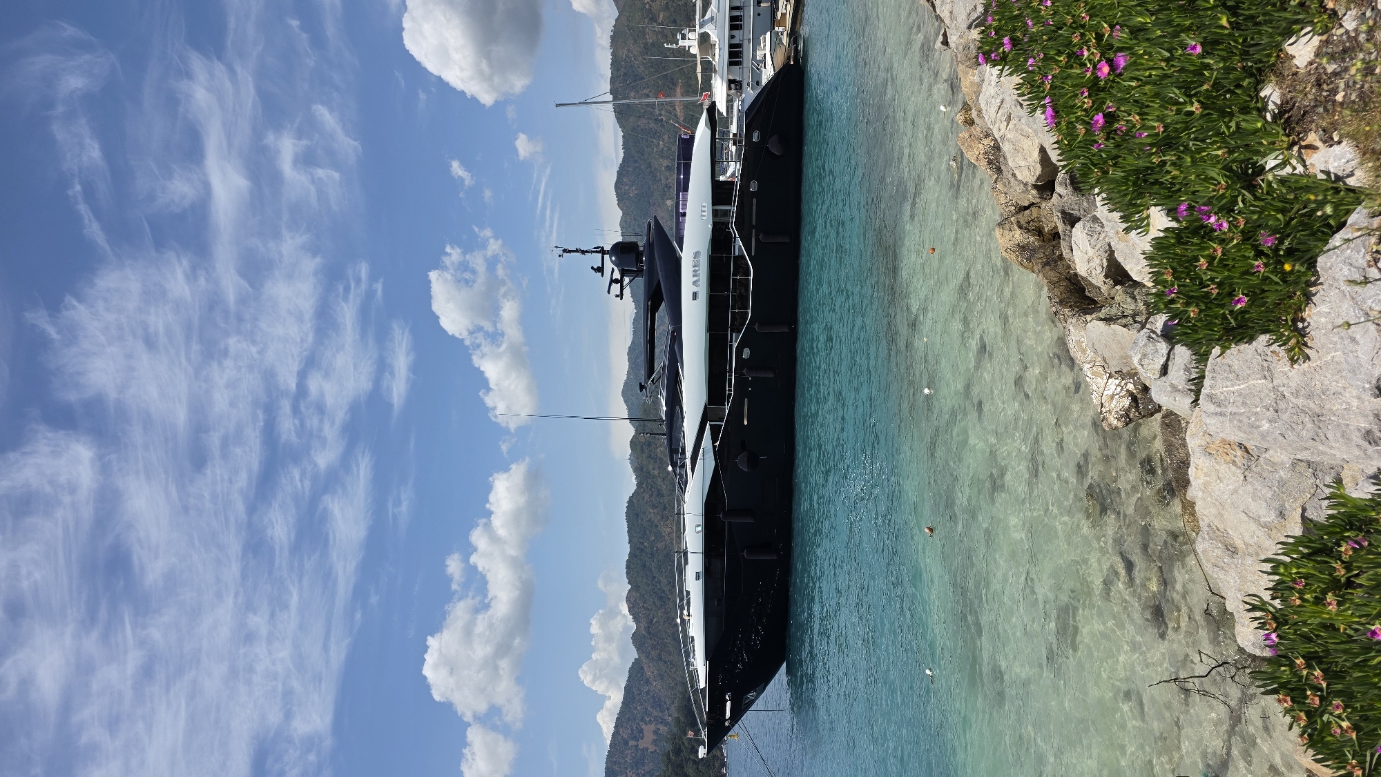 The image displays a serene coastal scene, likely in Fethiye, Turkey, under a clear, sunny sky. A prominent, modern yacht named "ARES" is the central subject, identifiable by the white text clearly visible on its dark hull. The yacht features a sleek black and white design with several decks and antenna masts, appearing to be docked or moored close to the shore.
The water is a striking turquoise-blue, exceptionally clear, and calm, reflecting the bright sky. Along the right side of the frame, a rocky coastline is visible, covered with vibrant green vegetation interspersed with numerous bright pink and purple flowers, possibly ice plants. Several small white buoys can be seen floating in the water near the shore.
In the background, a range of undulating mountains, covered in green foliage, forms the horizon beneath the sky. The sky itself is a brilliant blue, populated by a mix of delicate, wispy cirrus clouds and larger, fluffier cumulus clouds, indicating fair weather. The scene is devoid of visible human activity, conveying a sense of tranquility and natural beauty. The time of day appears to be bright daytime, likely late morning or early afternoon, given the clear illumination.