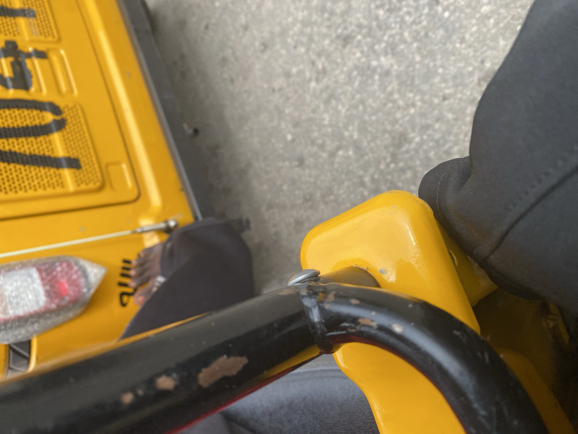 The image shows a close-up perspective from within a yellow auto-rickshaw in Jalingo, Nigeria. A person's dark-colored pants and feet are visible in the foreground, resting on the vehicle's floor. A black metal bar, part of the auto-rickshaw's structure, runs diagonally across the frame, showing some wear and tear. Behind the person's feet, the rear of the auto-rickshaw is visible, including a red taillight and part of a yellow panel with black lettering that appears to be a license plate. The ground, likely a road surface, is visible in the background, showing a rough, grey texture.