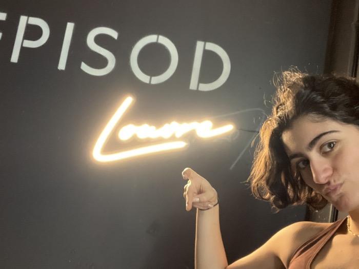 A young woman poses next to a wall sign. The sign reads "EPISOD" in a bold, stencil-style font above a cursive neon light spelling out "Laura". She is pointing with one hand at the neon sign. She has dark hair, brown eyes, and appears to be making a kissing face. She is wearing a tank top. The background is a dark wall.