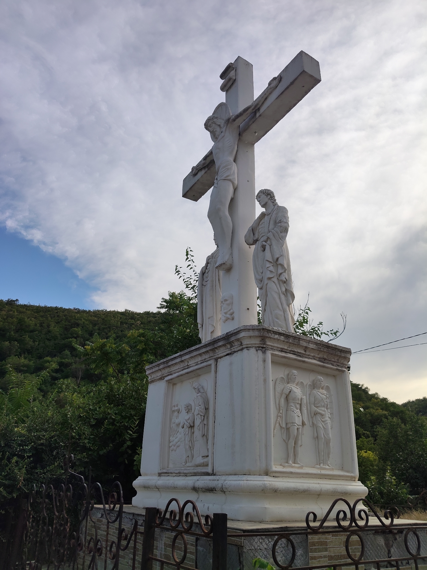 This is a low-angle, eye-level, full shot of a white stone crucifixion monument in Saharna, Moldova. The monument features a statue of Jesus Christ on the cross, with two additional figures at the base of the cross, one on the left and one on the right. The base of the monument is adorned with relief carvings of figures, including what appear to be angels and other religious figures. The monument is situated outdoors, with a lush green hillside covered in trees and vegetation in the background. The sky above is partly cloudy with patches of blue sky. In the foreground, a decorative metal fence with ornate scrollwork partially obscures the base of the monument. The overall scene depicts a religious monument in a natural, hilly environment.