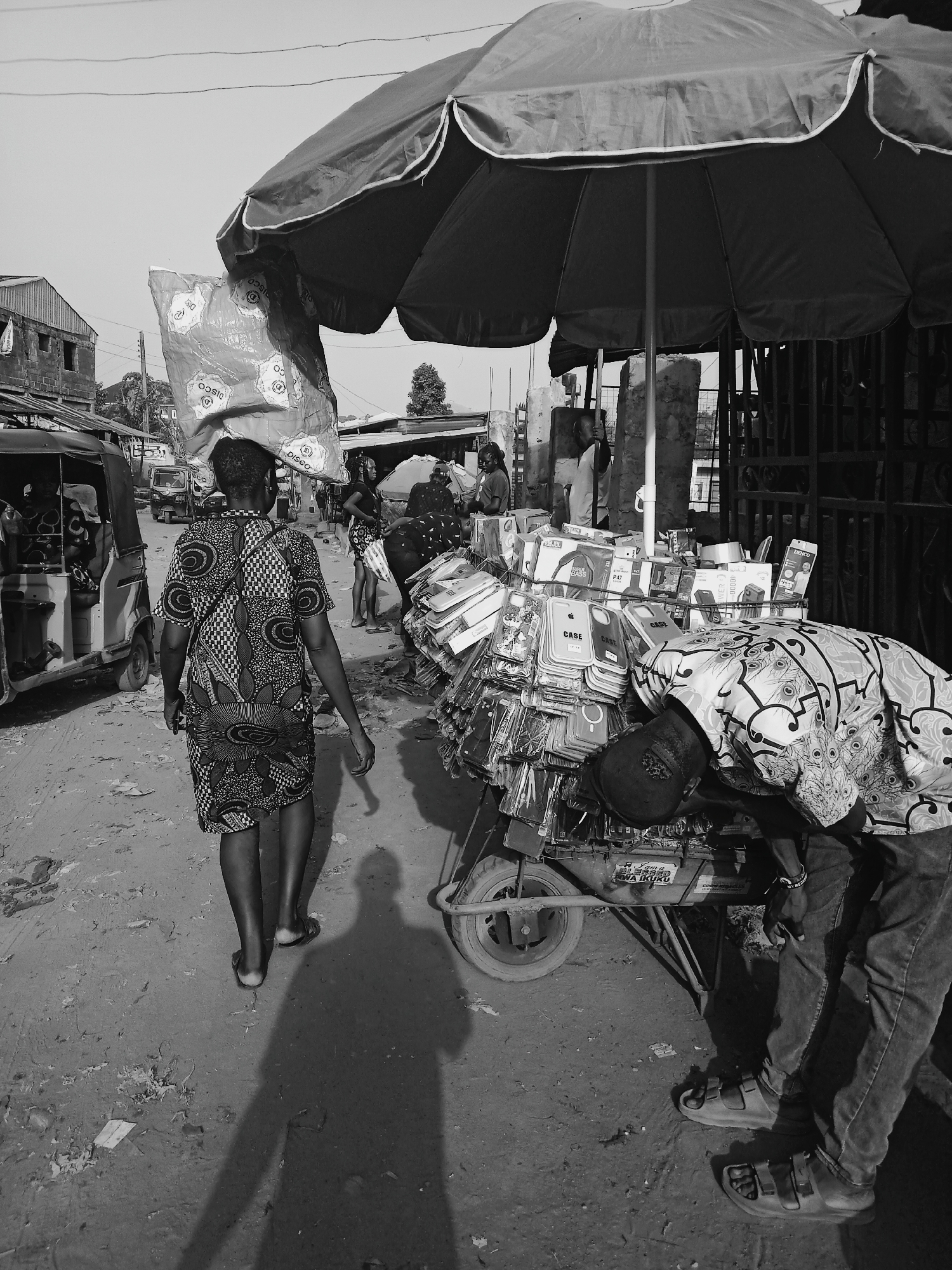 The image depicts a street scene in Onitsha, Nigeria. A woman in a patterned dress is walking away from the camera, carrying a large bag on her head. She is passing a man who is bending over a cart overflowing with phone cases and accessories under a large umbrella. The cart appears to be a mobile vendor stand. Other people are in the background, and there is a three-wheeled vehicle on the left. The ground is dirt and littered. The scene is monochrome.