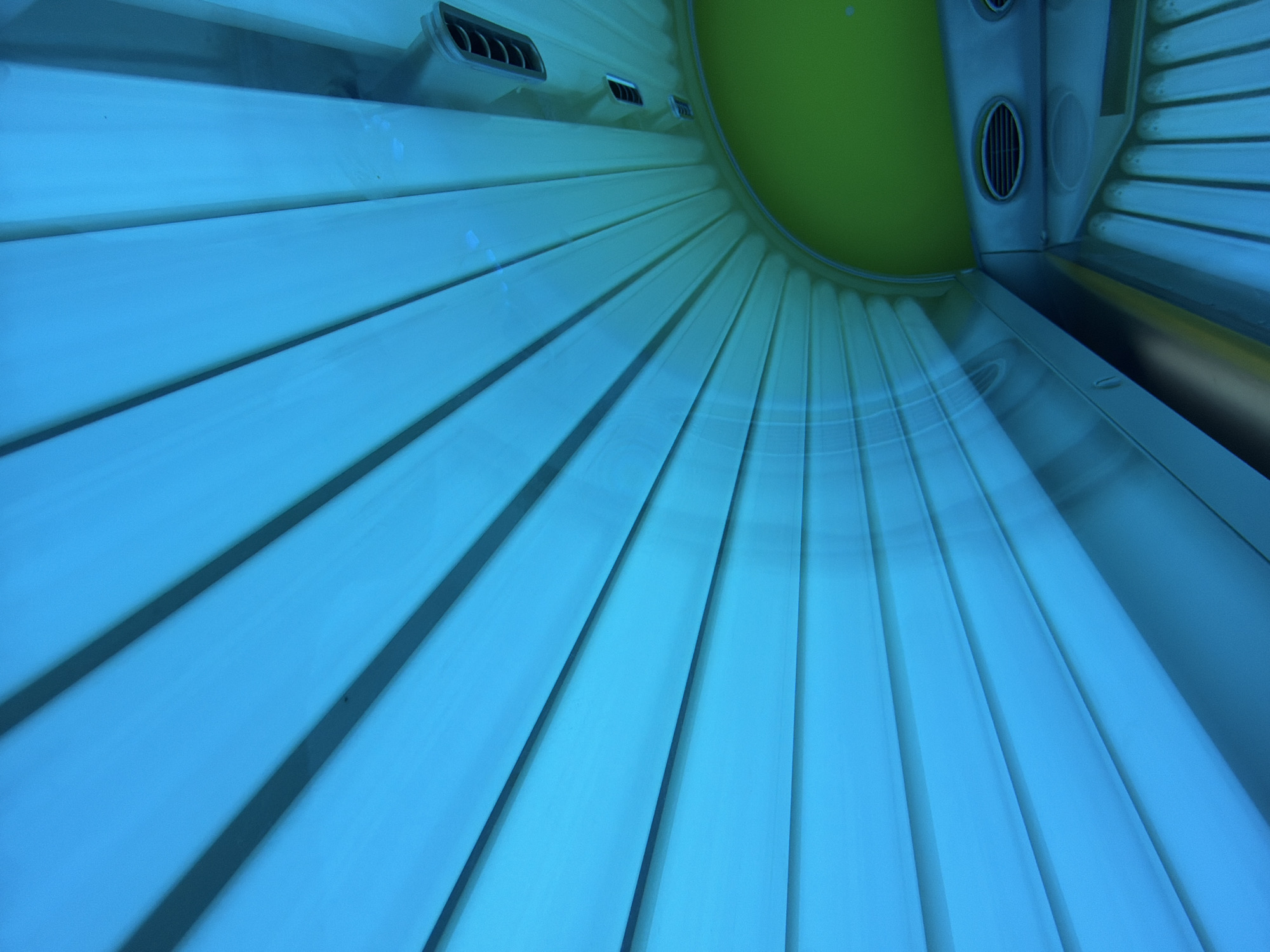 The image shows the interior of a tanning bed. The tanning bed is curved and lined with blue lights. There are also some green panels and ventilation grates visible. The image is taken from a low angle, looking up into the tanning bed. It is not possible to determine the location of this tanning bed within the city of Kyiv, Ukraine. There are no people or actions depicted in the image.