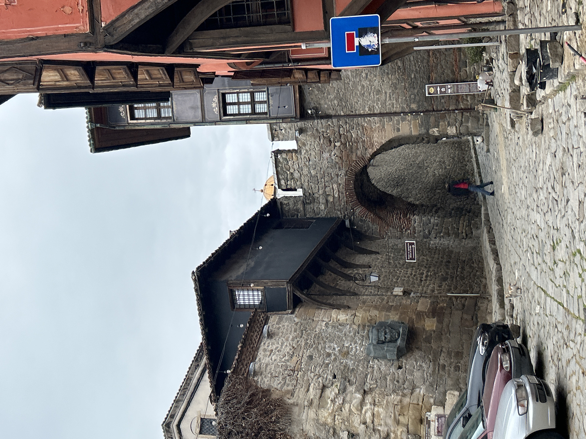 The image shows a street scene in Plovdiv, Bulgaria. The view is from a cobblestone street looking towards an archway. Buildings line both sides of the street, constructed of stone and wood, exhibiting traditional Bulgarian architectural styles. One building has an overhanging upper story supported by beams. A blue "dead end" street sign is visible. A person wearing a red jacket and jeans is walking through the archway. Cars are parked on the right side of the street. A stone carving is on the building near the arch. The sky is overcast. Construction equipment and debris are visible near the archway on the right side.