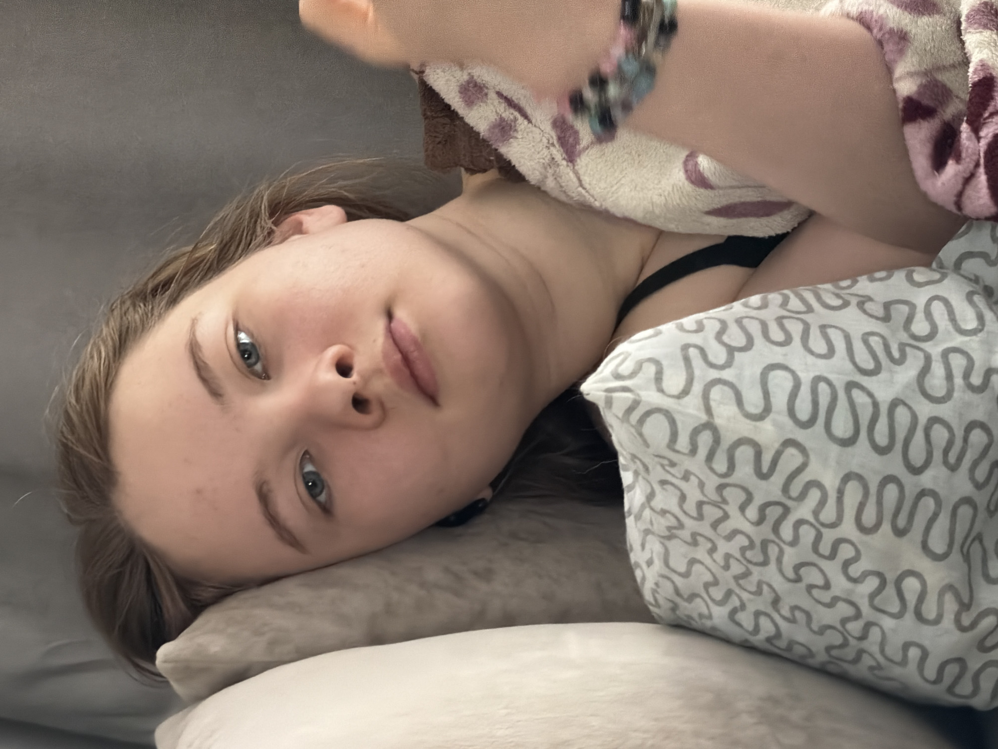 An image taken in the city of Chaikovsky, Russia, depicts a young female lying down indoors. The female, with light brown hair and blue eyes, is oriented on her left side, her head resting on a light-colored pillow. Her gaze is directed towards the camera. A black bra strap is visible on her left shoulder. Her right arm is partially visible, adorned with multiple beaded bracelets featuring blue, pink, and black beads. She is partially covered by a cream-colored blanket with purple and maroon patterns. A pillow with a white cover featuring a repeating gray serpentine pattern is positioned in the lower left foreground. The background consists of a plain, dark gray surface, likely a wall or headboard.