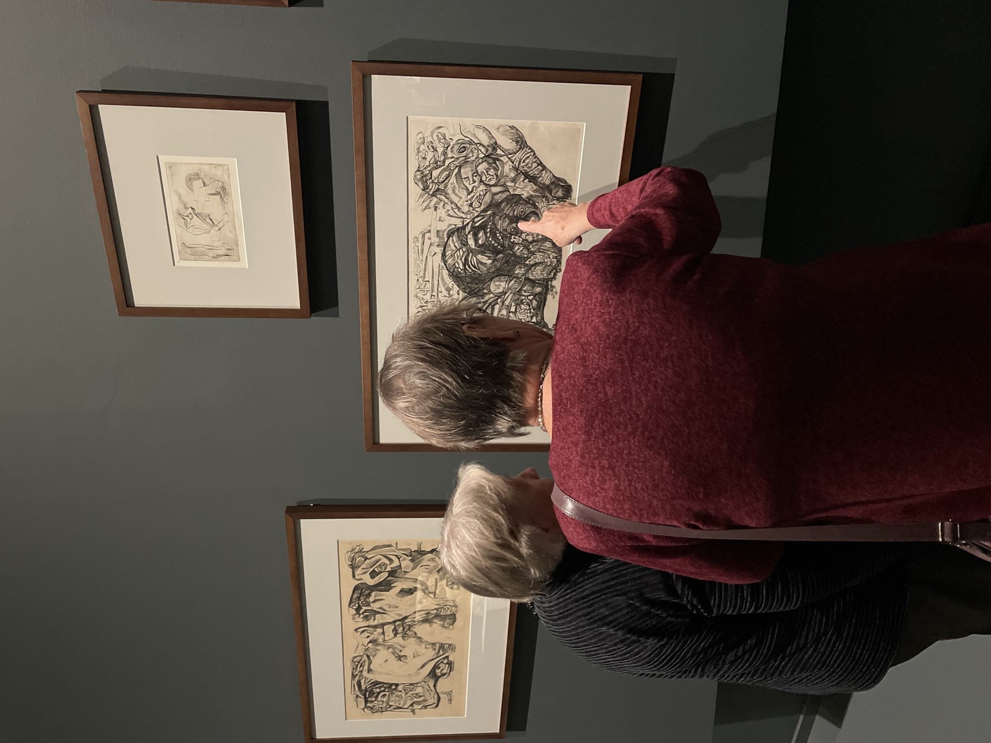 The image shows two adult females observing framed artworks within an exhibition space at the State Tretyakov Gallery on Krymsky Val, Moscow, Russia. The gallery wall is uniformly dark grey.
The woman on the left, with short light blonde or grey hair, is seen from the upper back and right profile, wearing a dark, possibly black or dark blue, vertically pleated long-sleeved top. The woman on the right, also seen from the back, has short grey hair and wears a maroon-colored long-sleeved top. A brown shoulder bag strap is visible over her left shoulder, and a light-colored necklace is around her neck.
The woman on the right extends her right arm and index finger, pointing directly at a specific detail within a large, horizontally oriented framed drawing. This drawing, prominently centered in the frame, features a dense, chaotic composition of grotesque and distorted faces and figures rendered in dark lines on a lighter background. A large, screaming or snarling face with an open mouth is central, surrounded by other contorted forms.
To the immediate left of this artwork, a second large, horizontally oriented framed drawing depicts multiple abstract or heavily stylized human figures in a dynamic arrangement. A smaller, horizontally oriented framed drawing hangs above and slightly to the right of the central artwork, showing simpler, sketch-like figural outlines. All three artworks are set within white matting and framed with simple light brown wooden borders. The environment is an interior gallery space with focused lighting, casting subtle shadows beneath the frames on the dark grey wall.