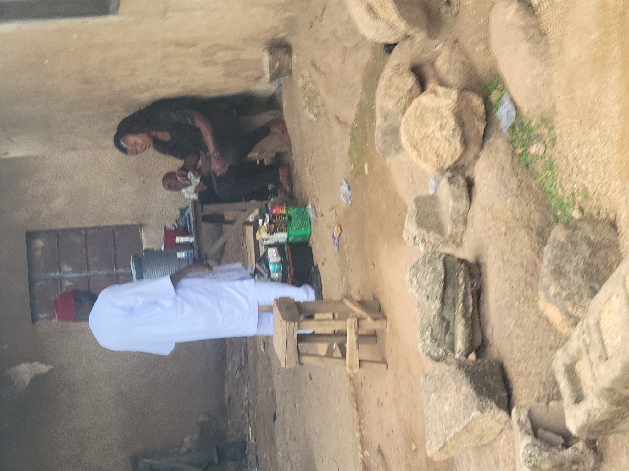 A man in a white robe and red cap stands facing away from the camera, his back to a rough-textured wall with a window. Beside him, a woman with long, dark, curly hair sits on a low bench or stool, holding a baby. She is wearing a black outfit. In front of the woman, a small table is laden with goods, including what appear to be bags of snacks or small items. To the left of the table is a green plastic basket filled with similar items. A wooden stool stands empty in the foreground. The ground is dusty and uneven, with several large, rough stones scattered around the scene. The overall environment suggests an outdoor market or informal settlement in the city of Jos, Nigeria.