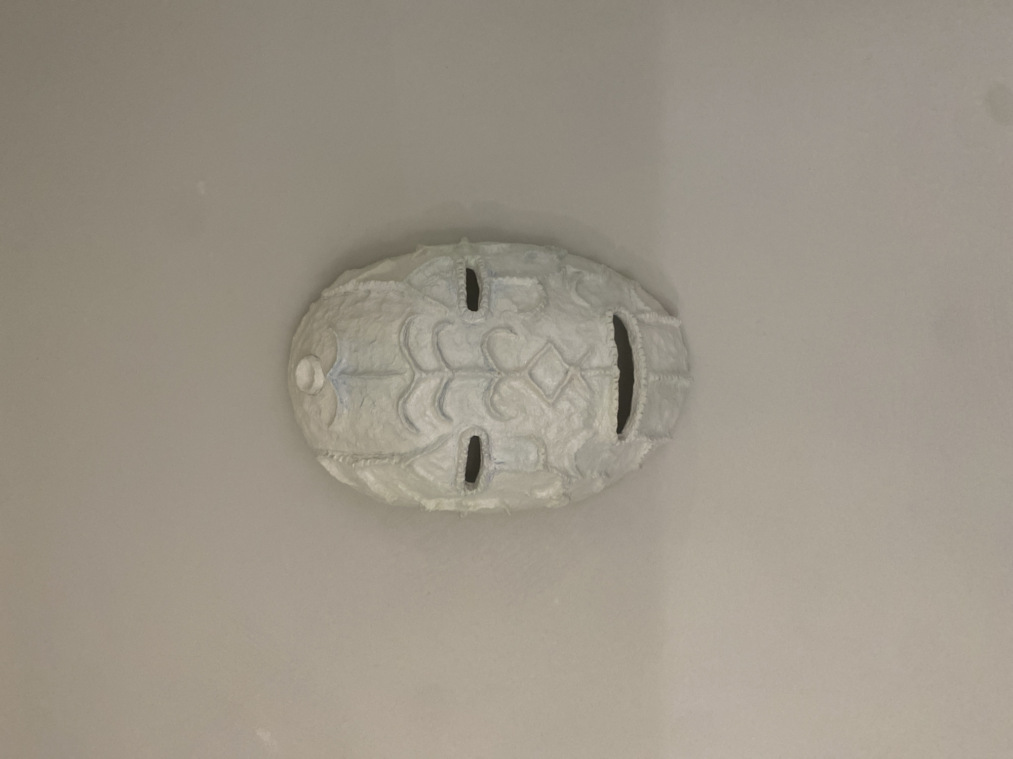 A white, possibly paper-mache mask is horizontally centered on a light gray surface. The mask has an oval shape with a central design suggestive of a stylized face. There are three dark oblong openings, one on each side and one towards the center of the mask. The surface is plain and light. The location information is not found in the image.