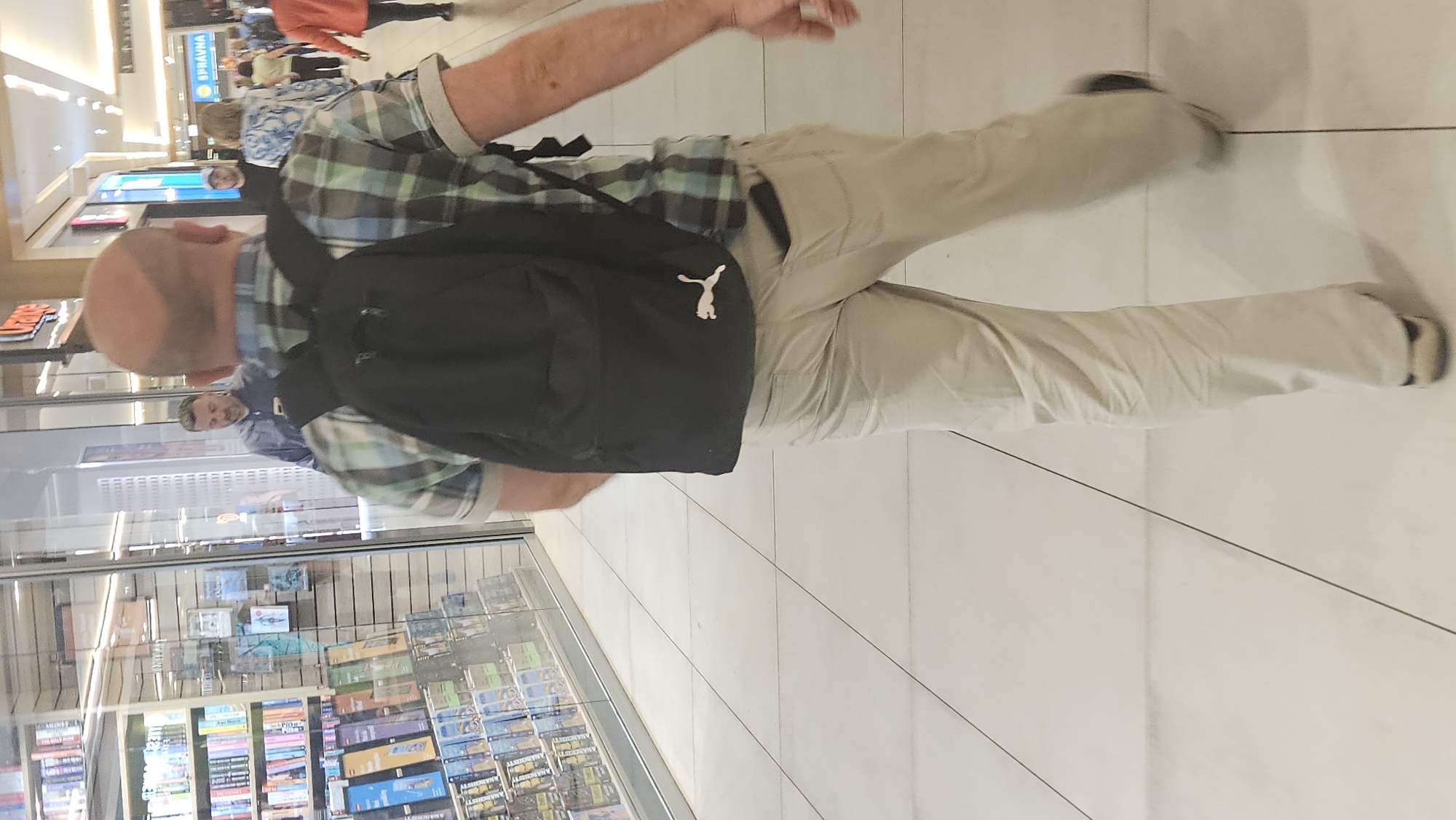 The image depicts the rear view of a bald man wearing a black backpack and khaki pants, walking in a brightly lit shopping mall. He is seen from the waist down, with his right arm slightly extended. The backpack has a white Puma logo visible on it. To his left, a bookstore display with numerous books is visible through a glass facade. In the background, other shoppers are seen walking in the mall, including a man in a blue shirt, a woman in a patterned top, and a person in an orange jacket. The mall floor is tiled in a light color. The environment suggests an indoor commercial setting.