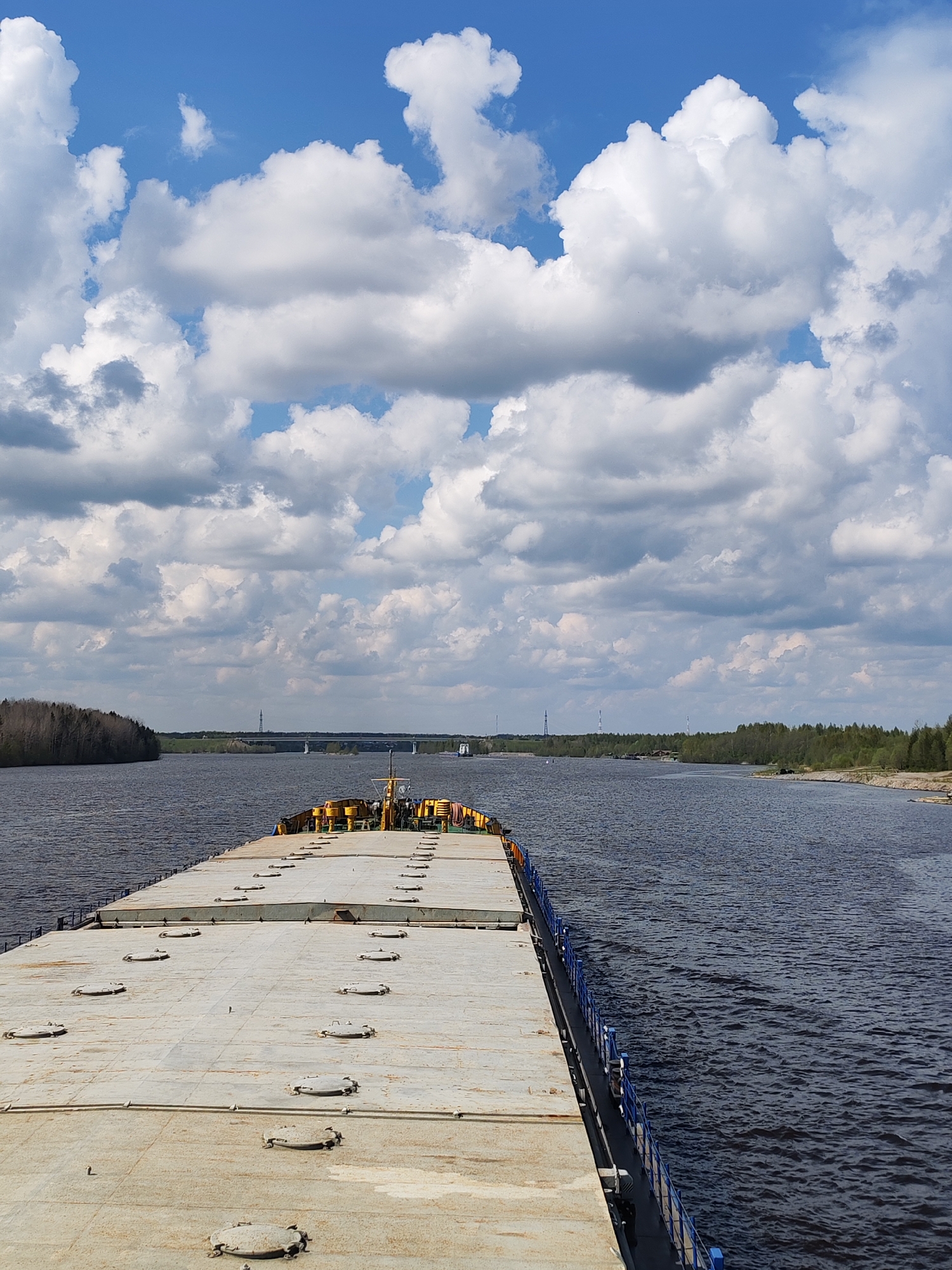 The image shows a barge traveling on a wide river under a partly cloudy sky. The barge is flat-topped with numerous hatches, and a yellow structure is visible at the far end. The river is flanked by green vegetation on both sides. A bridge and transmission towers can be seen in the distance. The location is unknown.