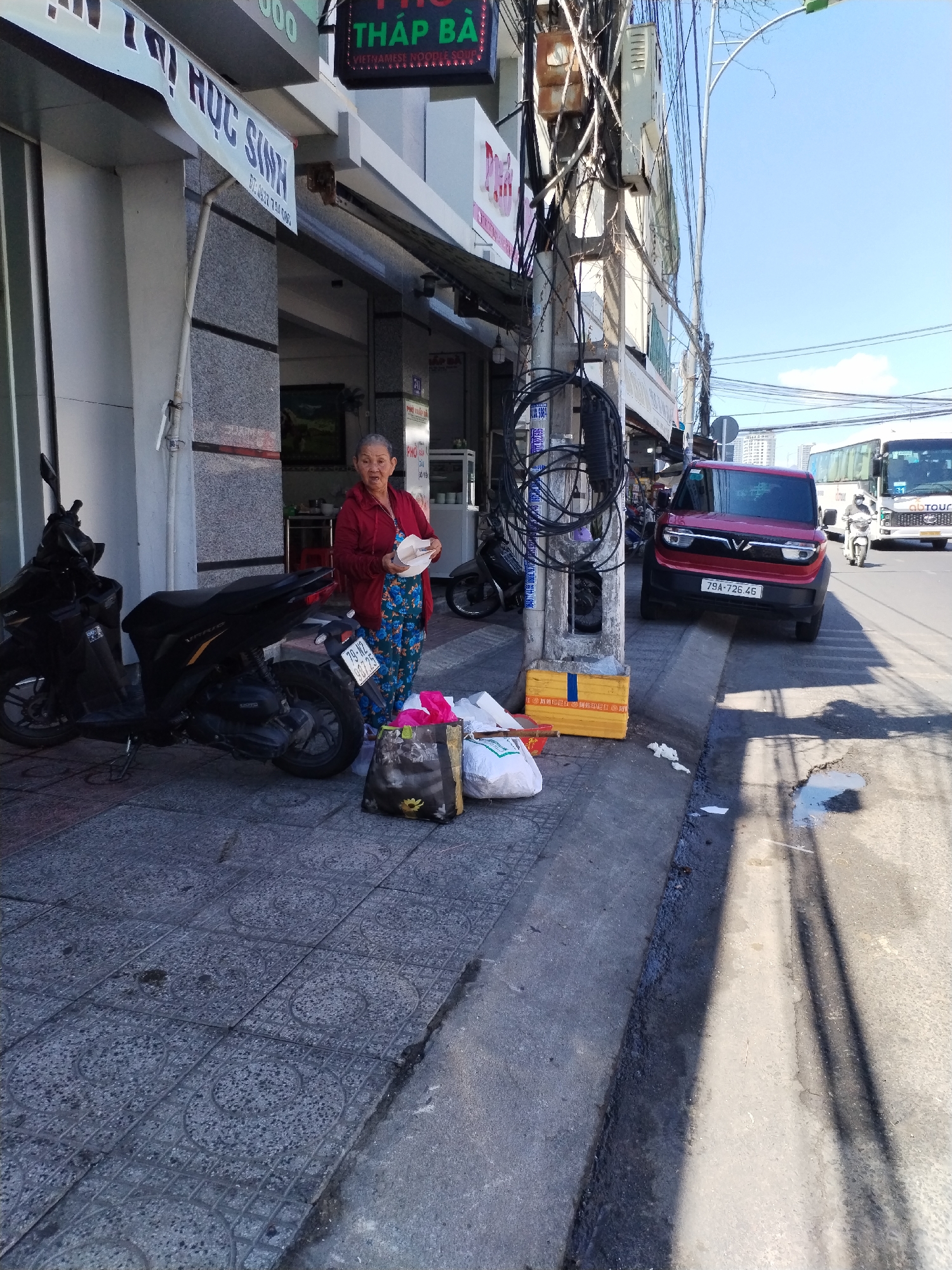 The image captures a street scene in Bac Nha Trang Ward, Vietnam. An elderly woman stands on the sidewalk, dressed in a red top and blue patterned pants, holding a white bowl. Several bags and a yellow crate sit near her. Parked behind her are two black motorcycles. To the right, a red car with license plate "79A-726.46" is partially visible, and a bus is further down the street. Overhead, a tangle of wires is connected to a utility pole. A sign reading "THAP BA VIETNAMESE NOODLE SOUP" is visible above the building, next to a sign "THI HỌC SINH." The sidewalk has a patterned tile surface, and the street is partially shaded.
