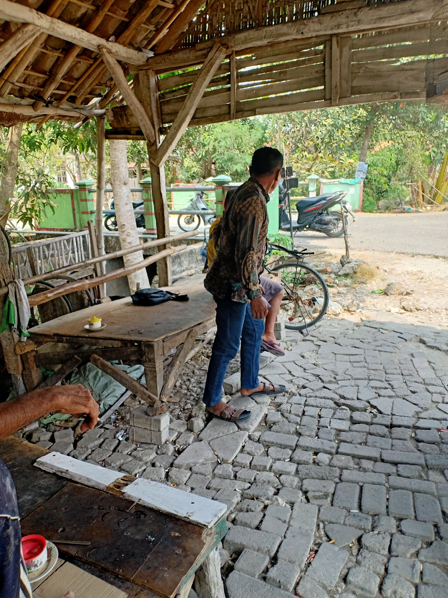 The image depicts an outdoor scene in Turigede, Indonesia, characterized by a rustic, open-air structure with a thatched roof supported by wooden beams. A man, wearing a patterned long-sleeved shirt and blue jeans, stands near a table and a bicycle. He is positioned with his back partially to the camera, looking towards the right. On the table, a small cup with a yellow lid sits on a saucer, alongside a black bag. The ground is paved with irregular stone blocks, some of which appear broken or uneven. Several cigarette butts are scattered on the ground between the paving stones. In the background, a road is visible, with motorcycles parked nearby, suggesting a public or commercial area. The environment is verdant, with trees and foliage contributing to a warm, sunny atmosphere. Another person's hand and arm are visible in the lower left corner, resting on a wooden surface, possibly indicating another individual present in the scene.