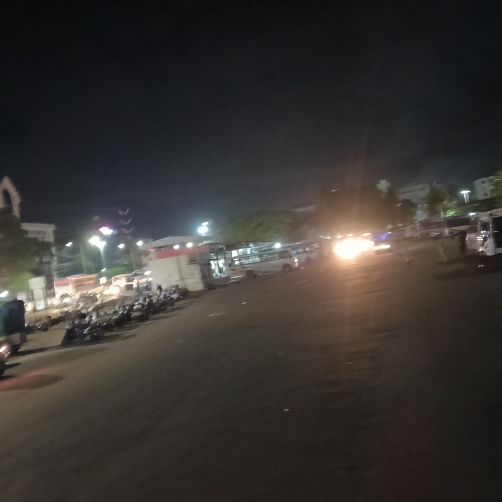 At night, a dimly lit, expansive public space in Margao, India, possibly a bus or transportation hub, is visible. A row of parked motorbikes lines the left side of the scene. A structure painted with red accents is present in the mid-ground, and buses are parked nearby. Vehicle headlights are visible in the distance. In the right side of the scene, there are white vans or small buses. A large, dimly lit area occupies the foreground.