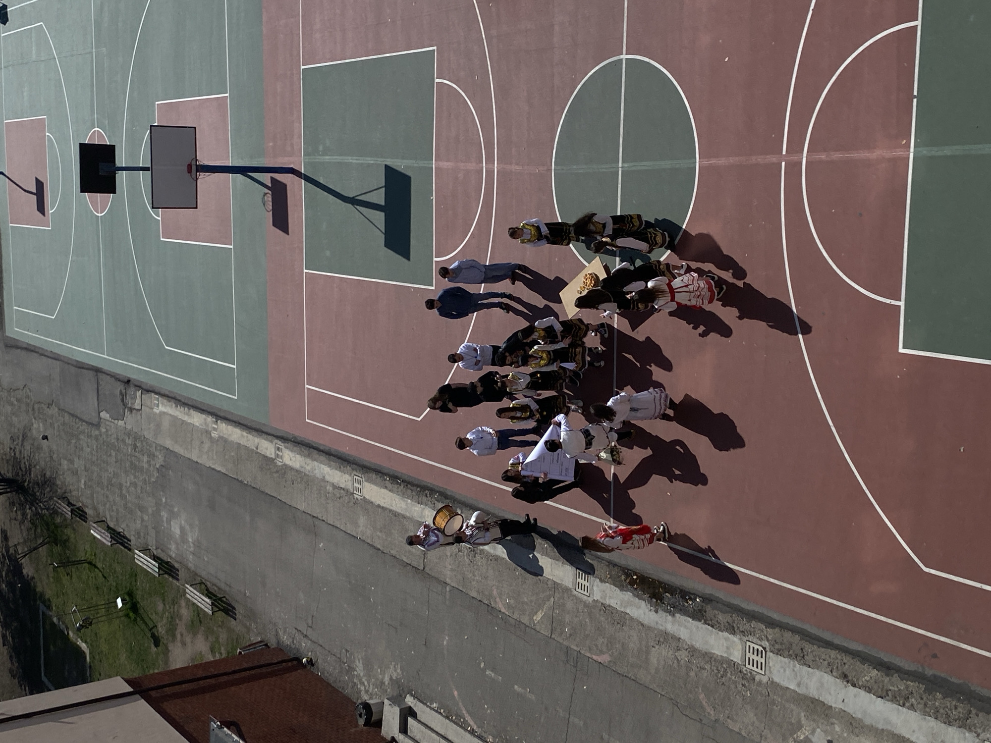 An overhead view captures an outdoor sports court, likely part of a school or public facility, situated in Sofia, Bulgaria. The court features a reddish-brown and green synthetic surface marked with white lines for multiple basketball games, including three visible basketball backboards and hoops. Strong midday sunlight casts prominent shadows from individuals and structures.
To the left of the court, a gray paved area separates it from a narrow strip of grass containing several wooden benches and sparse deciduous trees. A multi-story building with a red brick facade borders this area.
On the reddish-brown section of the court, a group of approximately 20 individuals is gathered in a semi-circular formation. The majority, predominantly young females, are attired in traditional Bulgarian folk costumes, characterized by white embroidered blouses, dark vests, and long, decorated skirts. Several men wear casual modern clothing (jeans, shirts), while two men near the group's left flank wear traditional or ceremonial uniforms. One of these men is actively playing a large tapan drum, and another appears to be holding a wind instrument.
In the central area of the costumed group, a small, light-colored wooden table or stand displays a platter of baked goods, possibly round pastries. Several individuals are holding and partially unfurling a large white banner or poster, which displays text and graphics. The individuals in traditional dress are generally standing, facing inward towards the center of the gathering. The scene suggests a cultural performance, celebration, or official event.