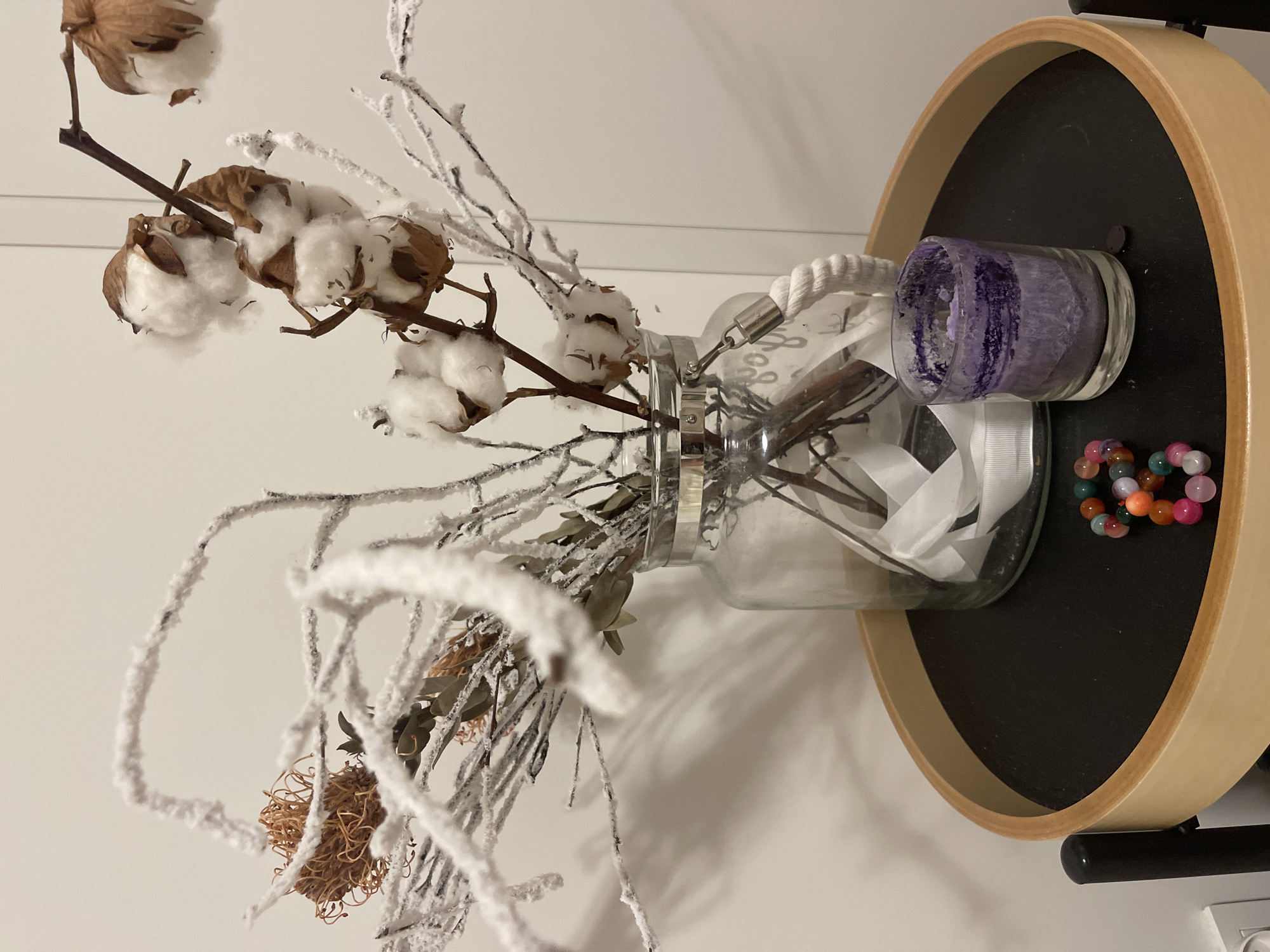 The image depicts a close-up of a decorative arrangement on a round, dark-surfaced table with a light wood rim. The arrangement includes a clear glass vase containing dried branches, some of which are dusted with a white substance resembling frost or snow. Among these branches are sprigs of cotton bolls, some appearing fully bloomed with fluffy white fibers, and others showing the brown husk. A piece of dried, brown, spiky flora is also visible within the vase. The vase itself has a silver band around its neck and a rope handle attached with a metal clasp resembling a lock. Inside the vase, white paper streamers are loosely placed.
In front of the vase, on the table, rests a small glass votive candle holder containing a partially melted purple candle. To the left of the candle is a bracelet or string of colorful beads, including shades of pink, orange, green, and white. The background is a plain, off-white wall.
The scene is set indoors, with artificial lighting casting subtle shadows on the wall to the left of the arrangement. No specific location in Wrocław, Poland, can be determined from the image. The image is a still life composition, focusing on the decorative objects.