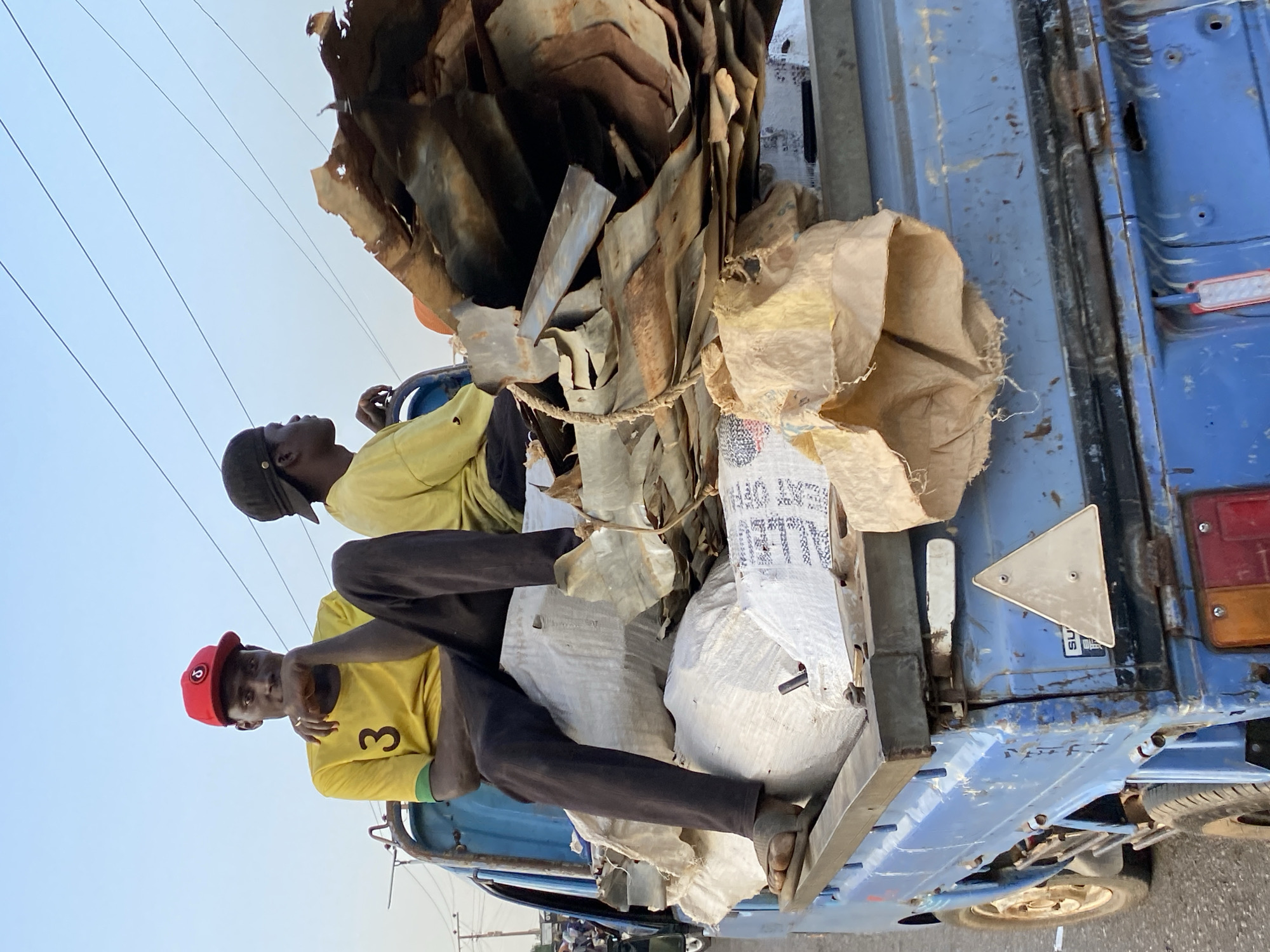 Two men sit atop the load of a small, open-backed blue truck. The truck bed is filled with a mixture of scrap metal, brown burlap sacks, and white sacks with black writing. The man in the foreground wears a red cap and a yellow t-shirt with the number "3". The other man wears a black cap and yellow t-shirt. The background contains overhead electrical lines. The location is Bukuru, Nigeria.