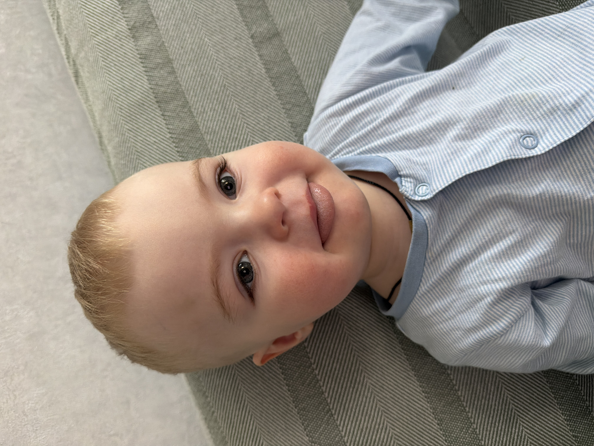 A close-up shot shows a baby lying on a striped, gray-green surface, likely a blanket or cushion. The baby is fair-skinned with blond hair, visible eyelashes, and is wearing a light blue and white striped shirt with buttons. The baby is looking up at the camera, smiling and sticking out its tongue. A thin black cord necklace is visible around the baby's neck. The background is slightly out of focus.
There is no information indicating this photograph was taken in Votkinsk, Russia.