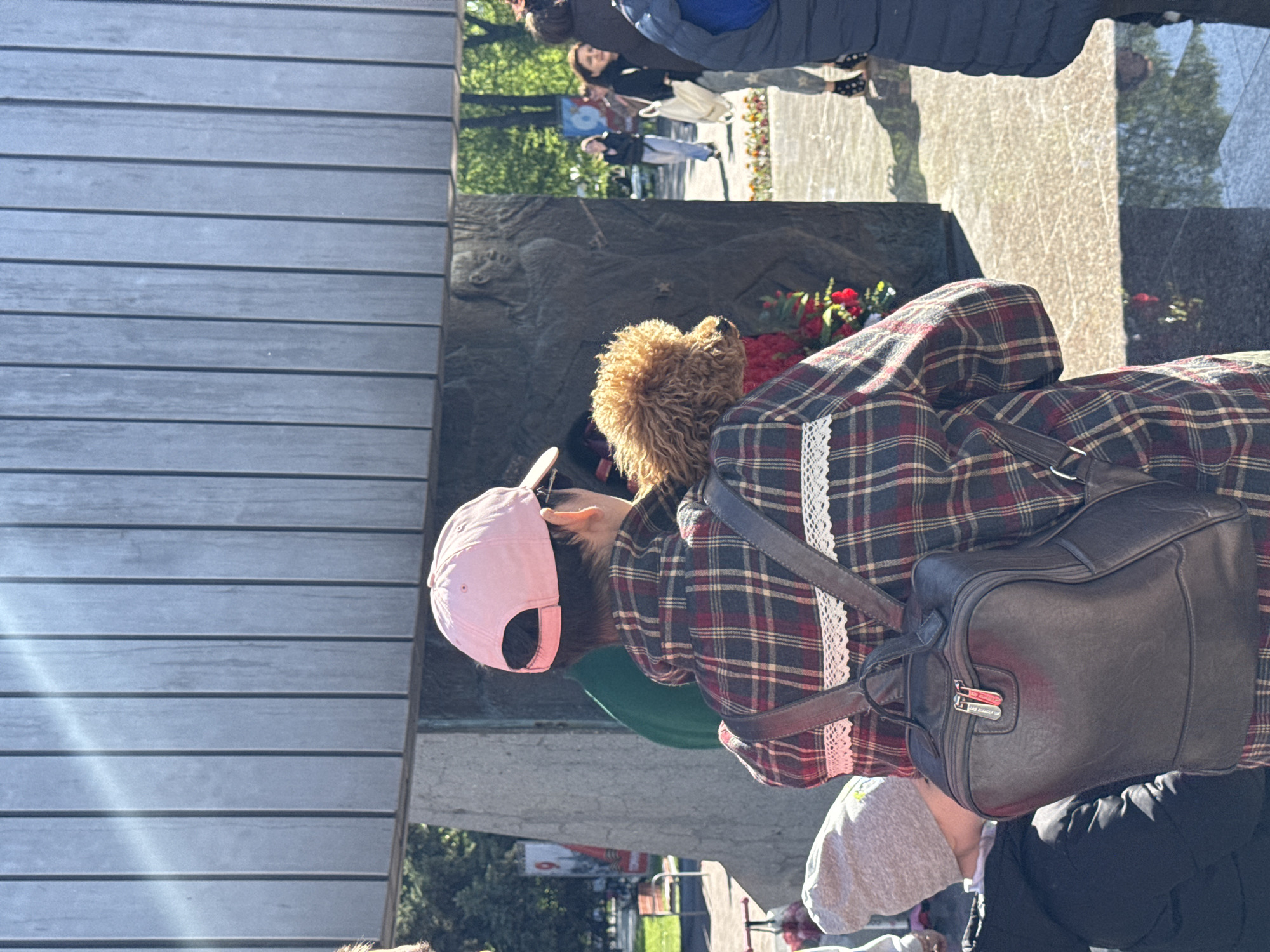 Here is a detailed description of the image:
The image shows a person wearing a pink baseball cap, a green shirt, and a plaid jacket with lace trim, standing at a war memorial. They are also wearing a backpack. A stuffed animal sits near the memorial. The memorial features a dark relief carving. People are visible in the background, some walking on a sidewalk. The scene appears to be outdoors in a city park. The location is Unknown, Unknown.