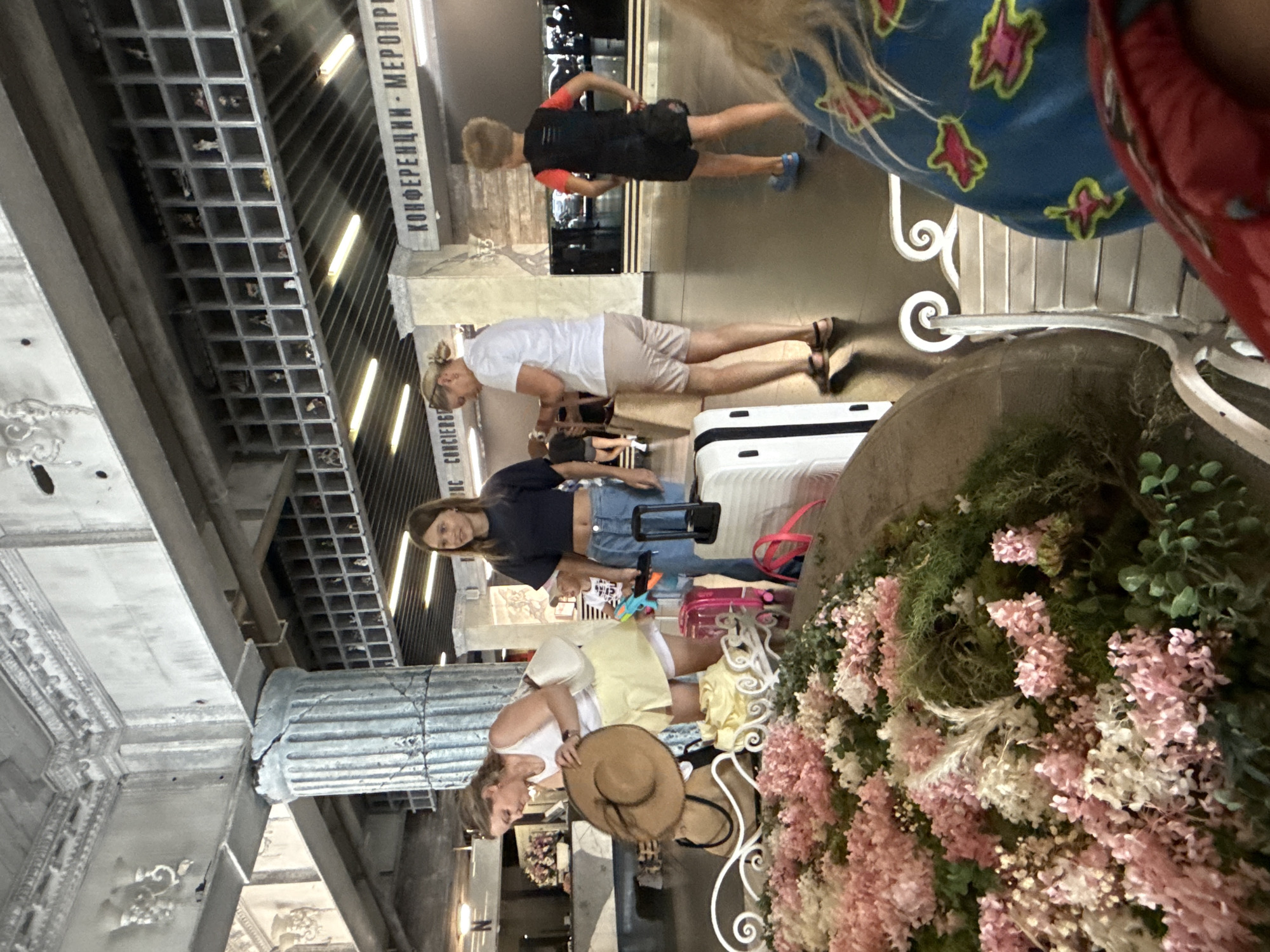 The image depicts an indoor scene, likely a hotel lobby or a similar public space, potentially in Yalta, Ukraine, given the Russian signage in the background that reads "КОНФЕРЕНЦИИ - МЕРОПРИЯТИЯ" (Conferences - Events). In the foreground, a large, decorative floral arrangement with pink and white flowers dominates the left side. Several people are visible in the mid-ground. A woman in a white top and yellow skirt is examining a straw hat, with luggage nearby. To her right, a teenage girl in a dark top and jeans stands looking to the right. A young boy in a black and red shirt and shorts is also present, facing away from the camera. The environment features classical architectural elements, including a large, fluted column with visible damage and ornate ceiling details. Shelving units filled with small figurines line the upper walls. The flooring is dark and reflective. The overall impression is that of a busy transition area, possibly a hotel lobby or a waiting area.