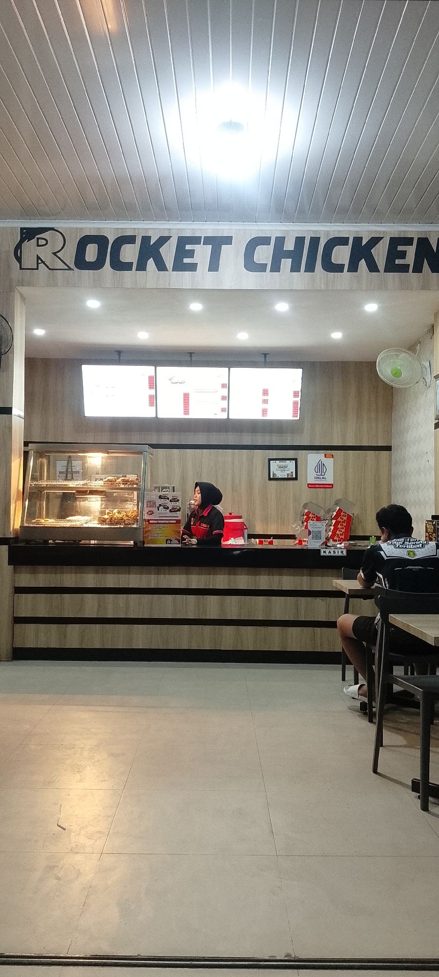 The image shows the interior of a "Rocket Chicken" restaurant in Batulicin, Indonesia. A female employee wearing a black hijab and a red and black uniform is standing behind a counter, attending to customers. In front of the counter, a male customer is seated at a table, facing away from the camera. The restaurant features a modern interior with wood-paneled walls and a suspended ceiling with fluorescent lighting. A display case showcases fried chicken, and menus are displayed on illuminated boards above the counter. The overall scene depicts a typical fast-food dining experience.