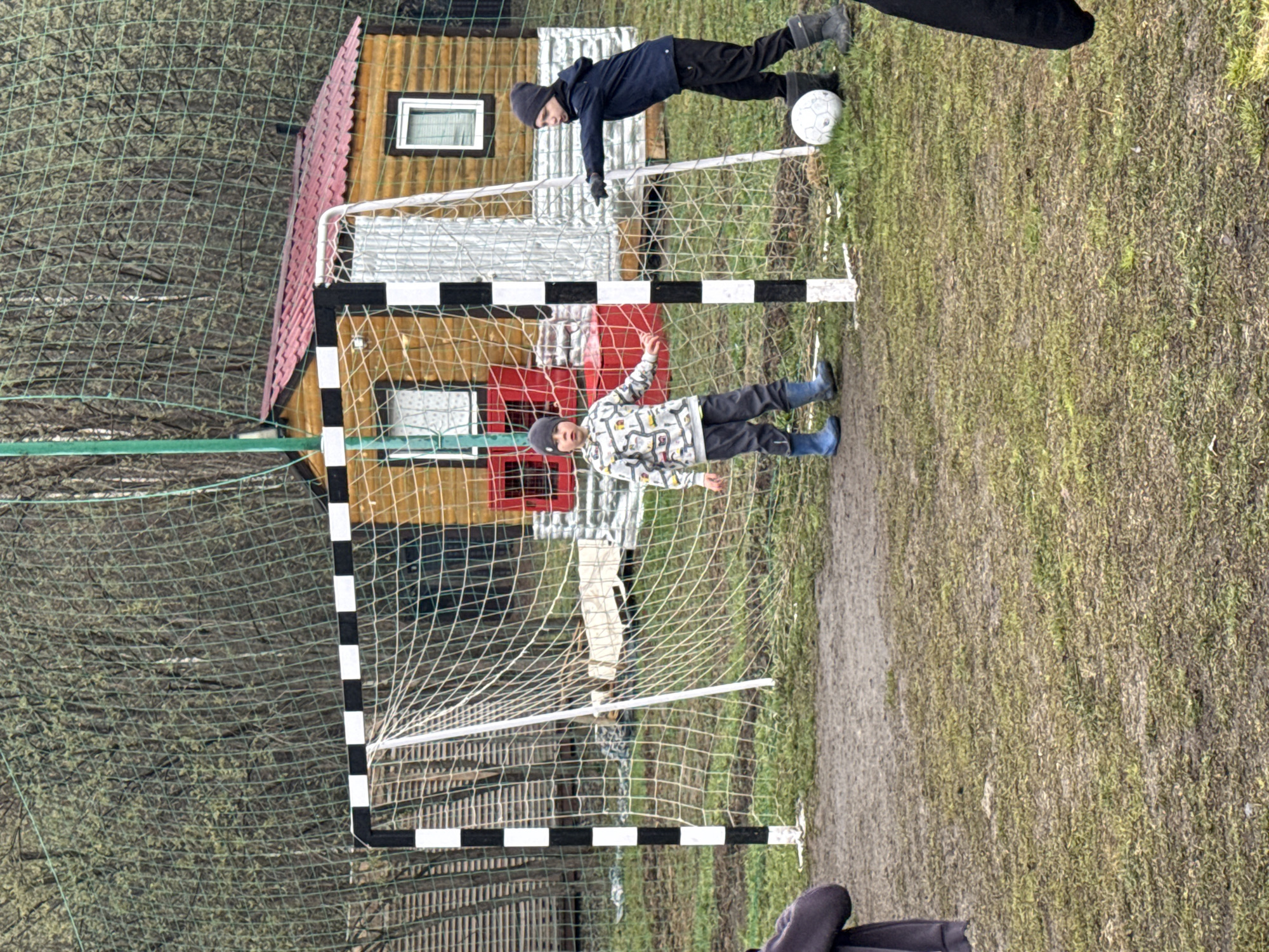 The image captures an outdoor scene where two children are engaged in playing soccer near a goal. In the foreground, on the right side of a soccer goal, a child wearing a dark blue jacket, black pants, black boots, and a dark beanie stands with a white soccer ball near their foot, appearing to be in motion or preparing to kick. Inside the goal, another child, dressed in a patterned white and grey jacket, light-colored pants, blue rubber boots, and a grey beanie, is positioned with arms slightly outstretched, resembling a goalkeeper. A partial view of an adult's legs and dark boots is visible in the bottom right corner of the frame.
The soccer goal itself features prominent black and white striped posts and a white net. To the left of the goal, in the mid-ground, stands a rustic wooden building or cabin with a reddish-brown tiled roof and windows. Behind this building and extending into the background is a dense area of bare trees, suggesting a wooded landscape, indicative of late autumn or early spring. The ground is a mixture of muddy dirt and sparse, dull green grass, with a visible dirt path running along the right side of the playing area.
The scene takes place during the daytime, likely an overcast day given the diffused lighting and lack of harsh shadows. The children's warm clothing, including hats and boots, suggests cool or cold weather conditions in Yekaterinburg, Russia. The overall mood conveys active, casual play. There is no visible text in the image.