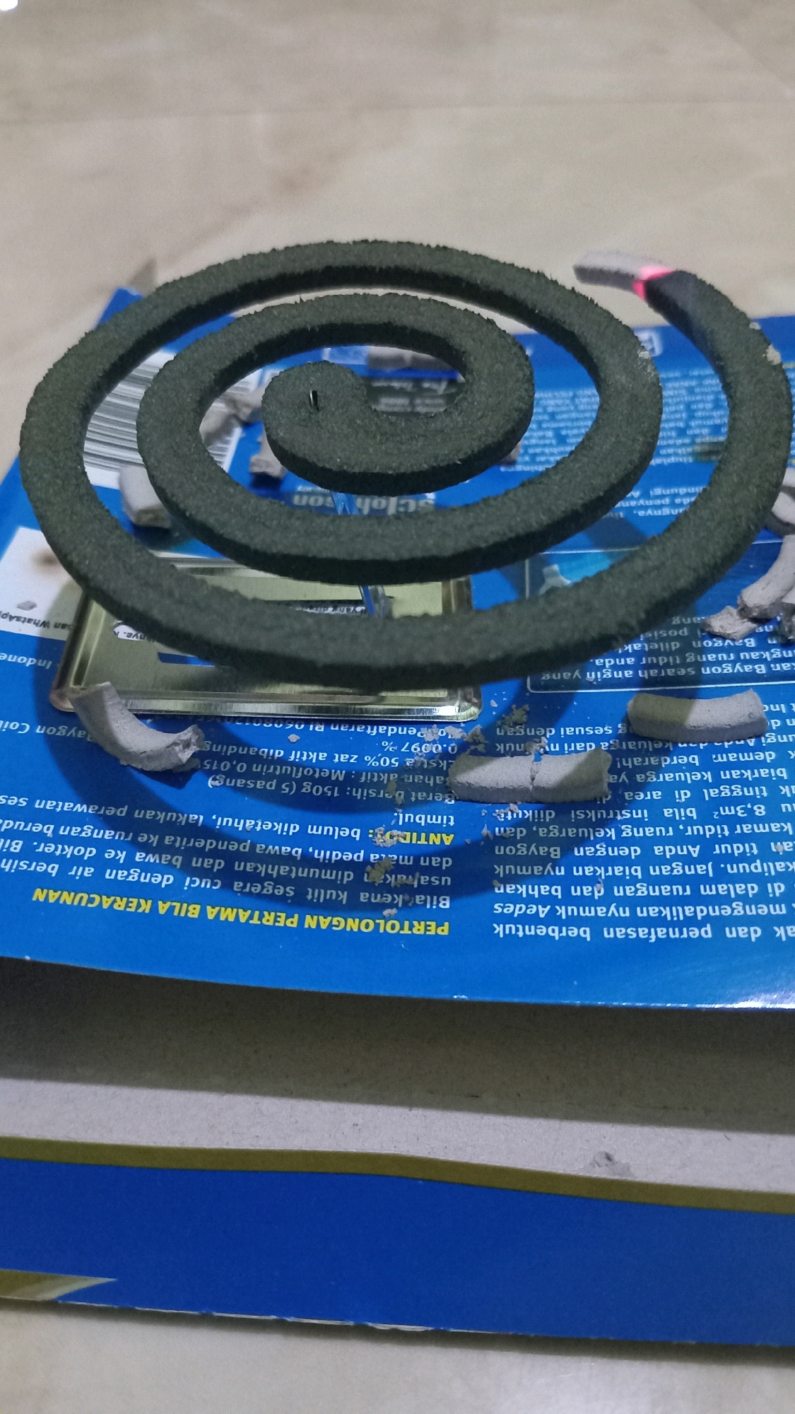 A close-up, low-angle shot shows a green mosquito coil placed on a blue cardboard surface. The coil is partially lit and emits a pinkish glow at its tip. Several small, broken pieces of the mosquito coil are scattered around the main coil. The blue surface has white and black text printed on it, some of which is obscured by the coil. The background is out of focus. The image is set in Randuagung, Indonesia.