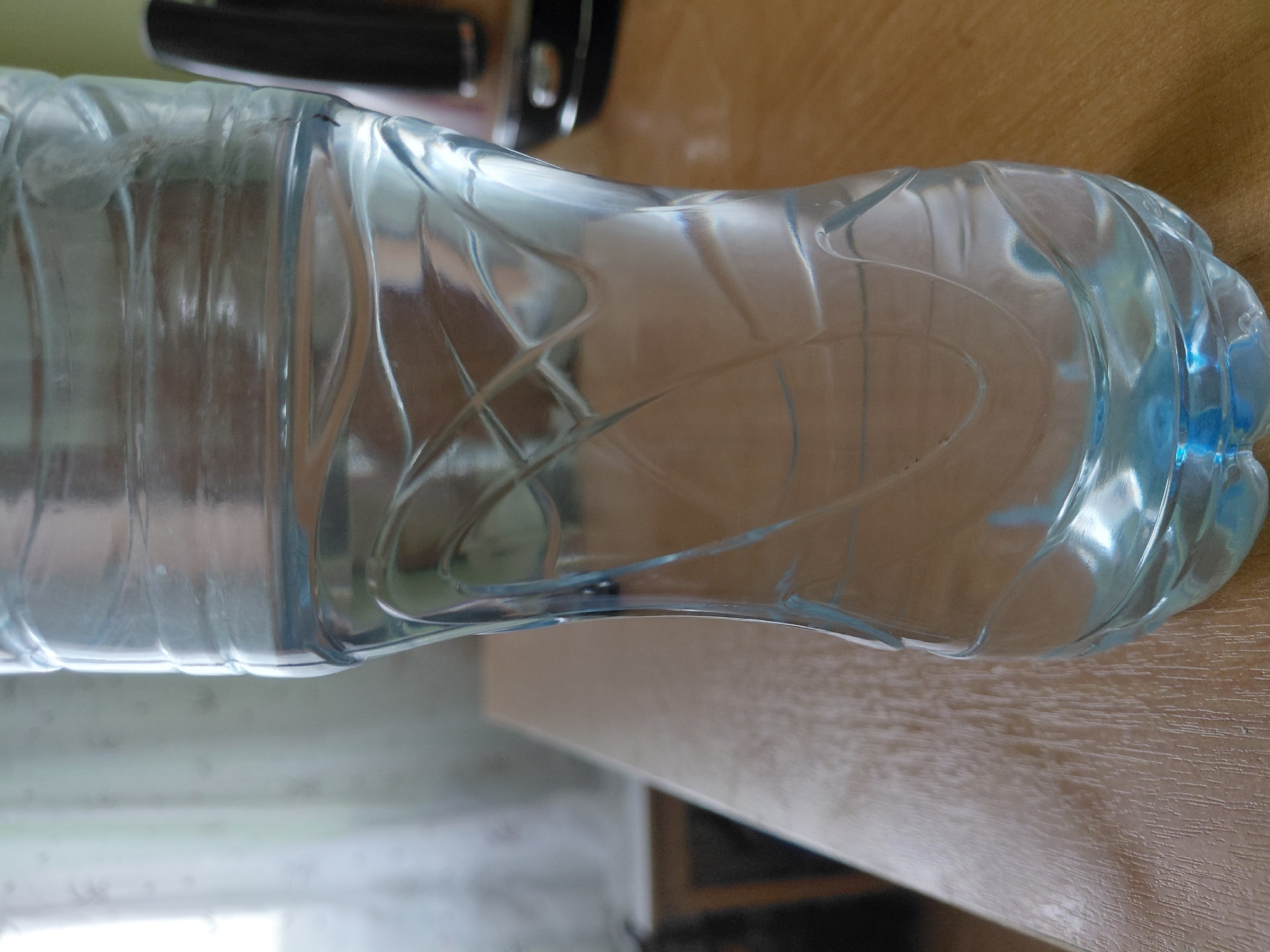 A plastic water bottle, partially filled with clear liquid, is the central subject of the image, lying horizontally across a light-colored wooden surface. The bottle is transparent with a slight blueish tint, particularly noticeable towards its cap end. Its body features a series of vertical ridges and interwoven, decorative patterns that create interesting refractions of light. The water inside appears still, with a subtle meniscus visible.
The bottle rests on a light brown wooden surface, likely a table or desk, which exhibits visible wood grain textures. In the blurred background, a dark object, possibly part of an appliance or furniture, can be seen, along with lighter areas that suggest a window or another light source. The lighting indicates it is daytime.
No specific activity or event is taking place; the image is a still life. No people are visible, and there are no discernible emotions conveyed. There is no visible text on the bottle or in the surrounding environment. While the location context provided is Nadym, Russia, there are no specific visual cues within the image itself to confirm this geographical detail.