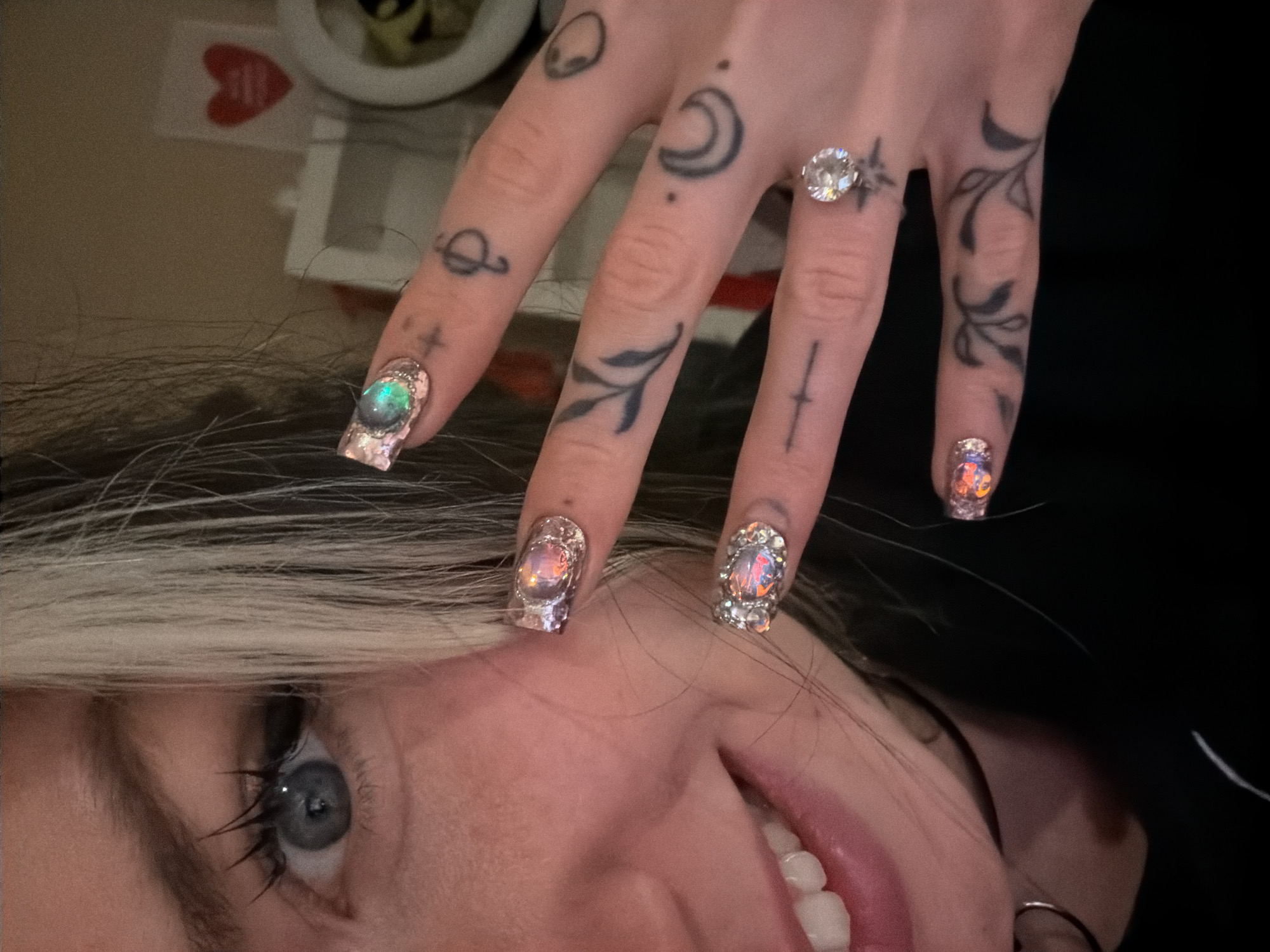 A close-up, rotated image captures a person's face and one hand, showcasing intricate details. The person has blonde hair falling across their forehead, bright blue eyes with dark mascara, and a gentle smile revealing white teeth. Their head is tilted significantly, appearing almost upside down in the frame.
The hand is adorned with numerous small, dark tattoos on the fingers and knuckles. These include designs such as an alien head, a stylized Saturn, a crescent moon with dots, leafy branches, a small cross, and a dagger or sword. A prominent, sparkling clear stone ring, possibly a diamond, is worn on the ring finger, surrounded by a star tattoo.
The fingernails are long and elaborately decorated. Each nail features a central, iridescent or opalescent cabochon-style embellishment that shimmers with various colors like green, purple, orange, and pink. These central stones are encircled by smaller, clear rhinestones or glitter, creating a highly decorative and reflective nail art design.
The background is softly blurred, suggesting an indoor setting. Hints of objects such as a red heart shape and a white circular item are vaguely visible. The overall impression is a personal photograph, possibly a selfie, focusing on the individual's distinctive style through their tattoos and ornate nails. No discernible text is visible within the image.