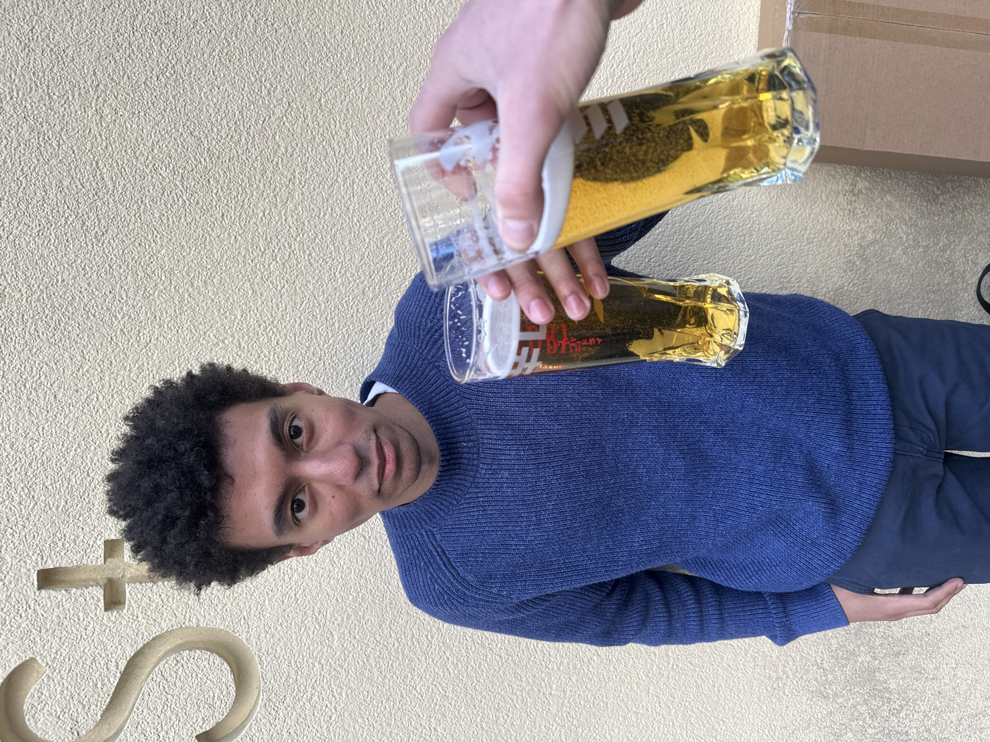 A man with dark curly hair wearing a blue sweater and dark pants is being photographed while holding a glass of beer as someone pours beer into it from another glass. The location appears to be outdoors against a textured light-colored wall. A partial metal "S" or decorative element is affixed to the wall, as well as a plus-shaped symbol. A cardboard box is visible in the upper right. The location is not London.