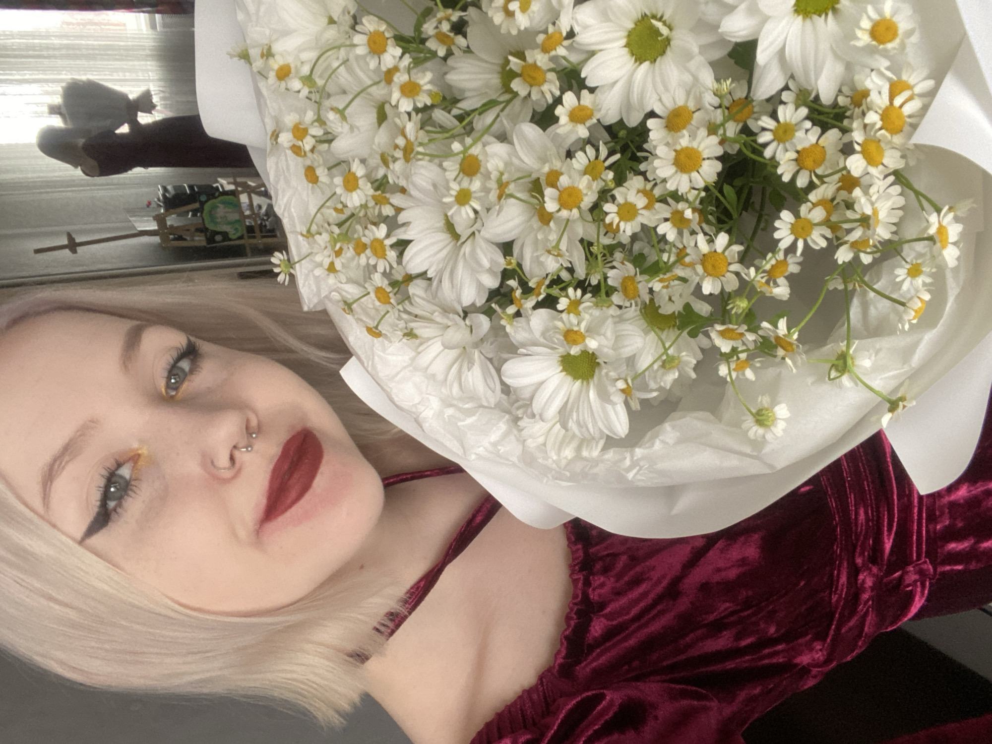 A young woman with blonde hair, light-colored eyes, and prominent winged black eyeliner is depicted holding a large bouquet of flowers. She has dark red lipstick and two visible nose piercings: a silver septum ring and a stud in her left nostril. She is wearing a dark red, possibly velvet, dress or top with thin straps.
The bouquet, filling much of the right side of the frame, is composed of numerous white daisies or similar daisy-like flowers, distinguished by their white petals and bright yellow centers. The flowers are wrapped in simple white paper.
The background is softly blurred, showing reflections in what appears to be a window. These reflections include indistinct shapes and a small, light-colored wooden object with green details, suggesting an indoor setting. The woman has a subtle smile, conveying a pleasant or happy expression. There is no visible text in the image. This media file is associated with the location context of Izhevsk, Russia.