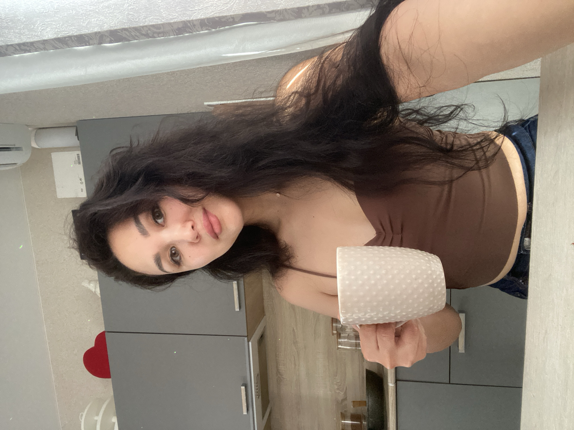 This media file captures a selfie of a young woman in an indoor setting, likely a kitchen or utility room. The woman has long, dark, wavy hair and a light complexion, looking directly at the camera with a neutral expression. She is wearing a brown spaghetti strap top, with a portion of blue jeans visible on her right side. She holds a white, textured mug in her right hand.
The background features dark grey kitchen cabinets with silver handles and a light-colored countertop. An air conditioning unit is mounted on the wall to the upper left. Below the air conditioner, a white paper or cardboard item with a barcode and indistinct text (including a copyright symbol) is visible. A patterned curtain or blind hangs above the air conditioner. A red heart-shaped object is positioned on the floor or a lower surface to the left. The scene appears to be well-lit, suggesting daytime. The image depicts a casual moment in a residential environment located in Vysokoye, Russia.