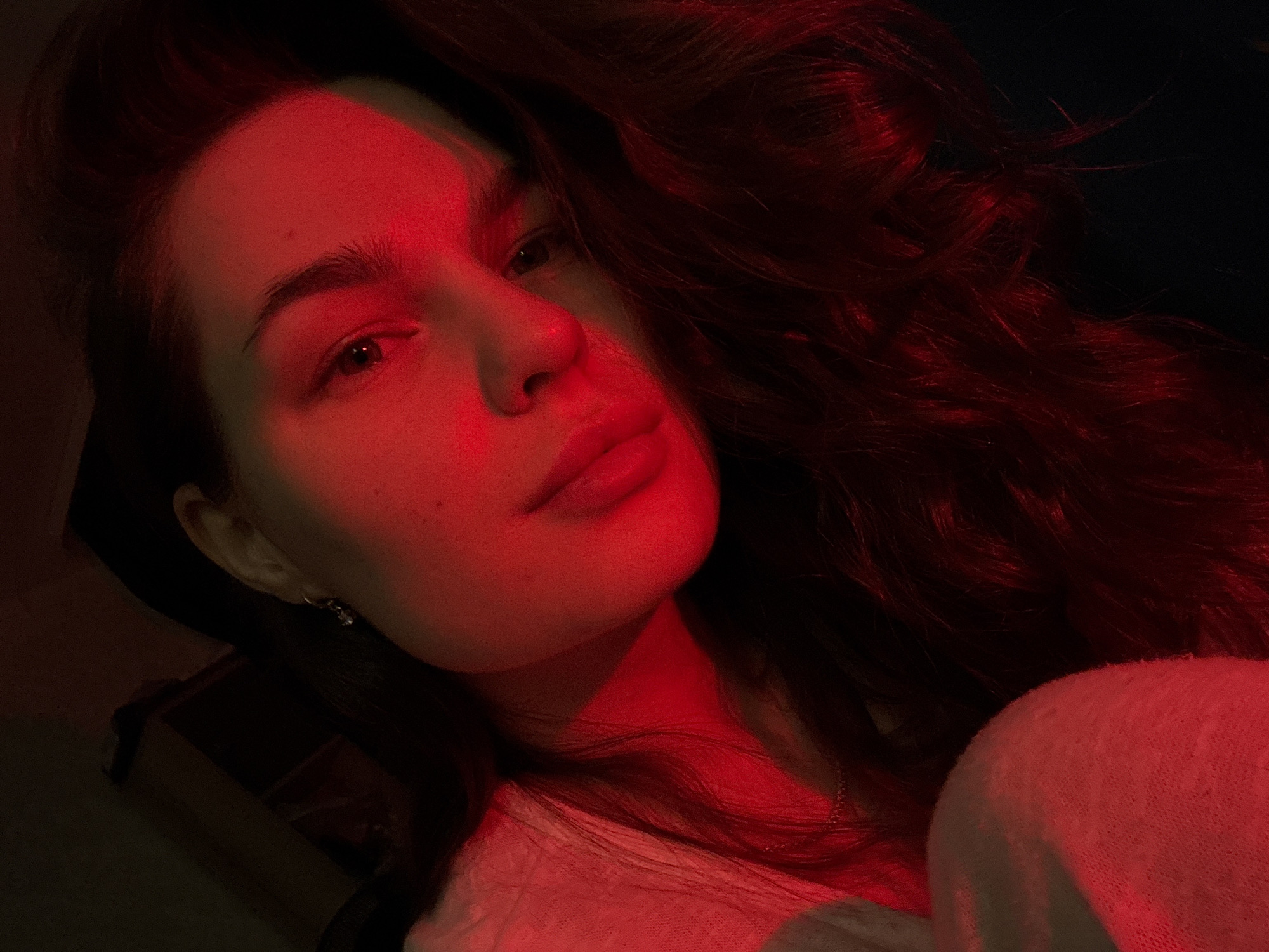 The image is a close-up, low-angle, self-portrait of a young woman illuminated by red light. She has long, dark, wavy hair and appears to be lying down, with her head resting on her shoulder. Her left cheek is prominent, and her eye is visible, looking off to the side. She is wearing a light-colored, textured garment. The background is dark and indistinct, suggesting an indoor setting. There is no discernible location in the city of Chaikovsky, Russia, visible in the image.